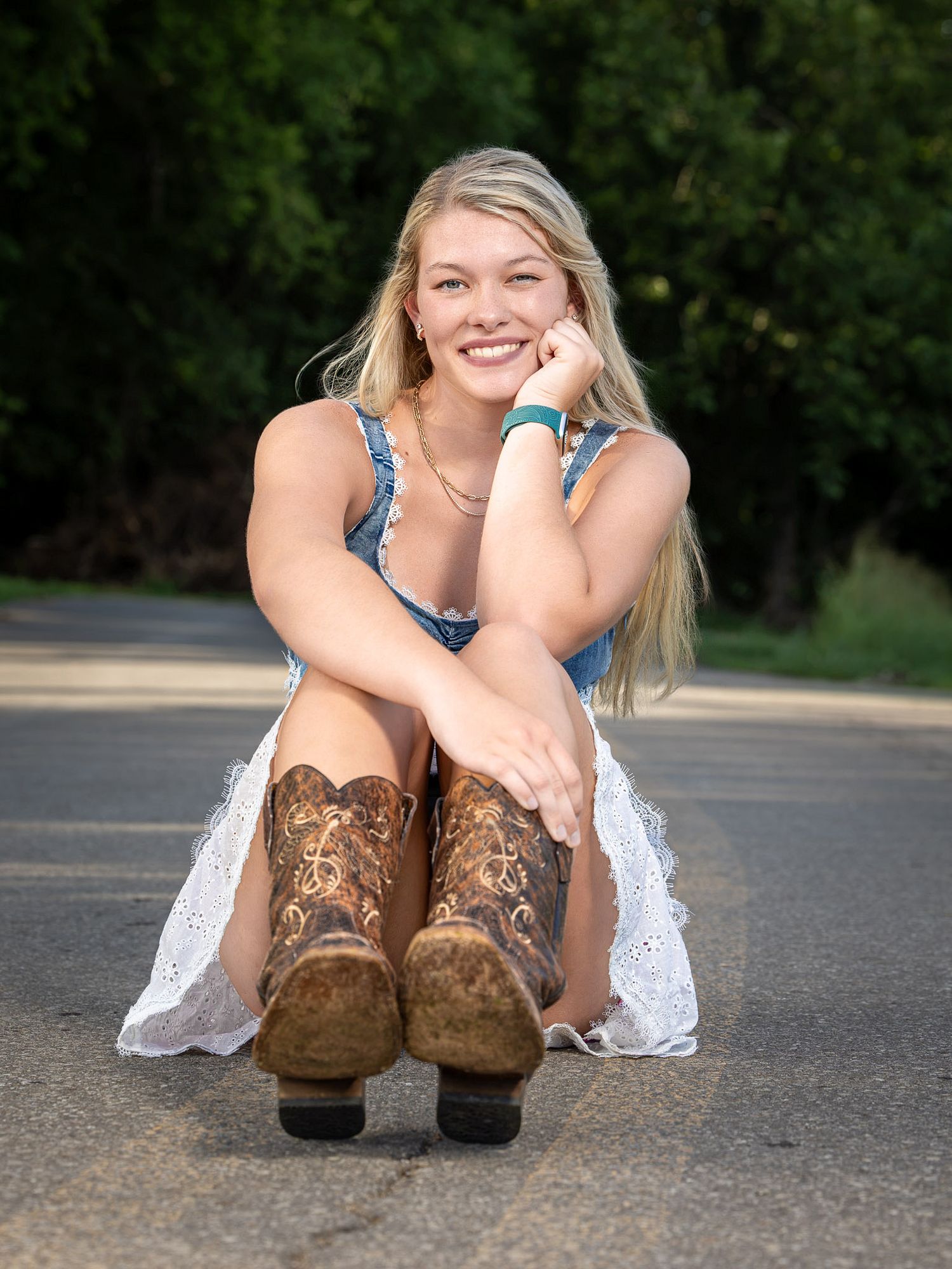 Smiling blonde woman sitting on a road wearing cowboy boots, a denim top, and a white lace skirt.