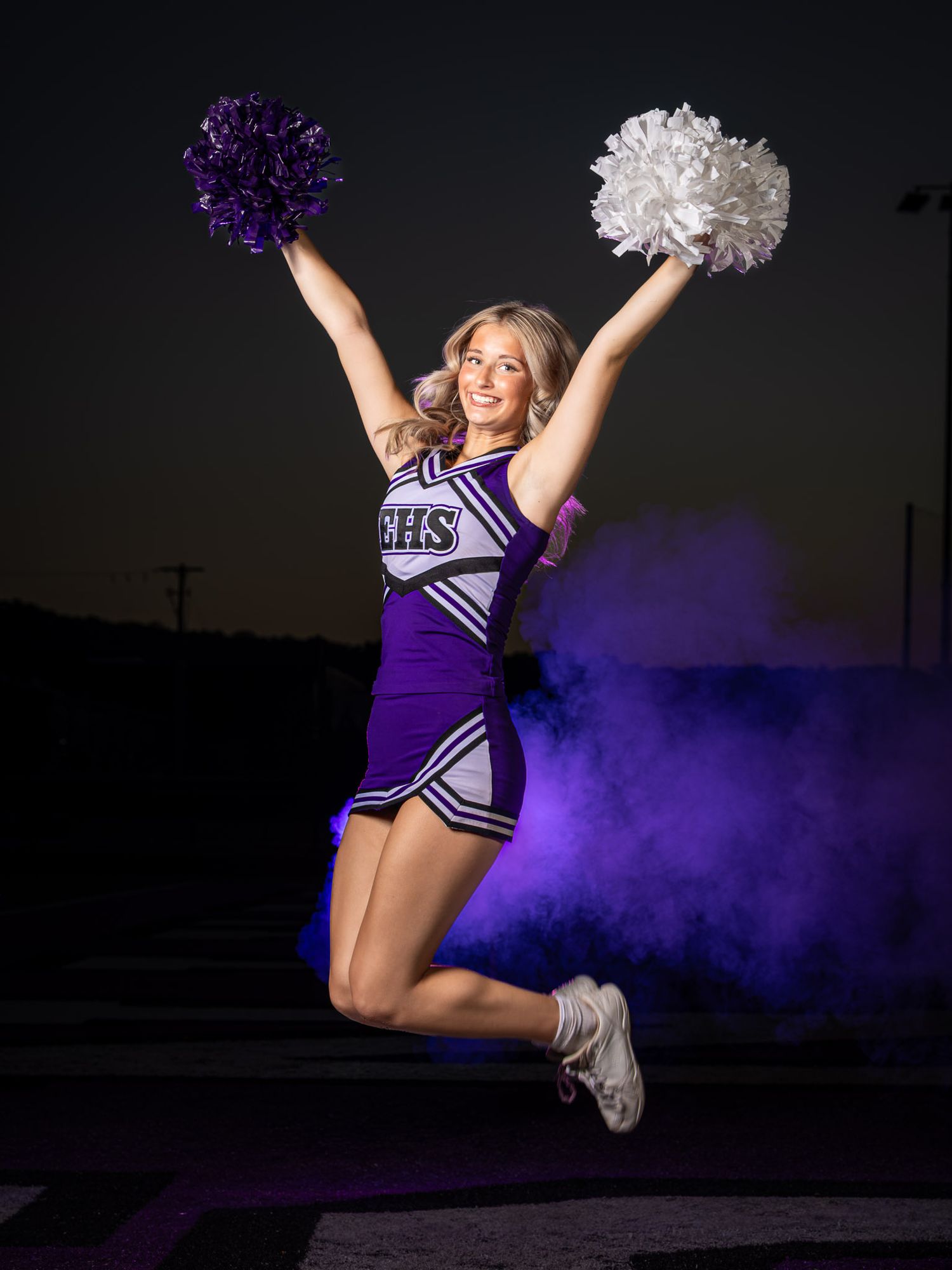 A cheerleader in a purple BHS uniform jumps mid-air holding purple and white pom-poms with dramatic smoke behind her.
