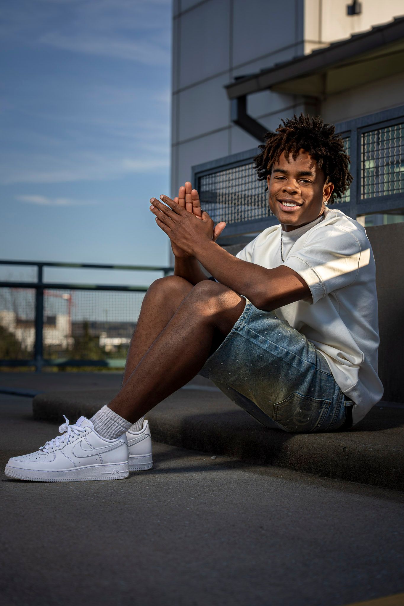 Smiling young Black man sitting outdoors in white tee, denim shorts, and Nike Air Force 1 sneakers, clapping hands.