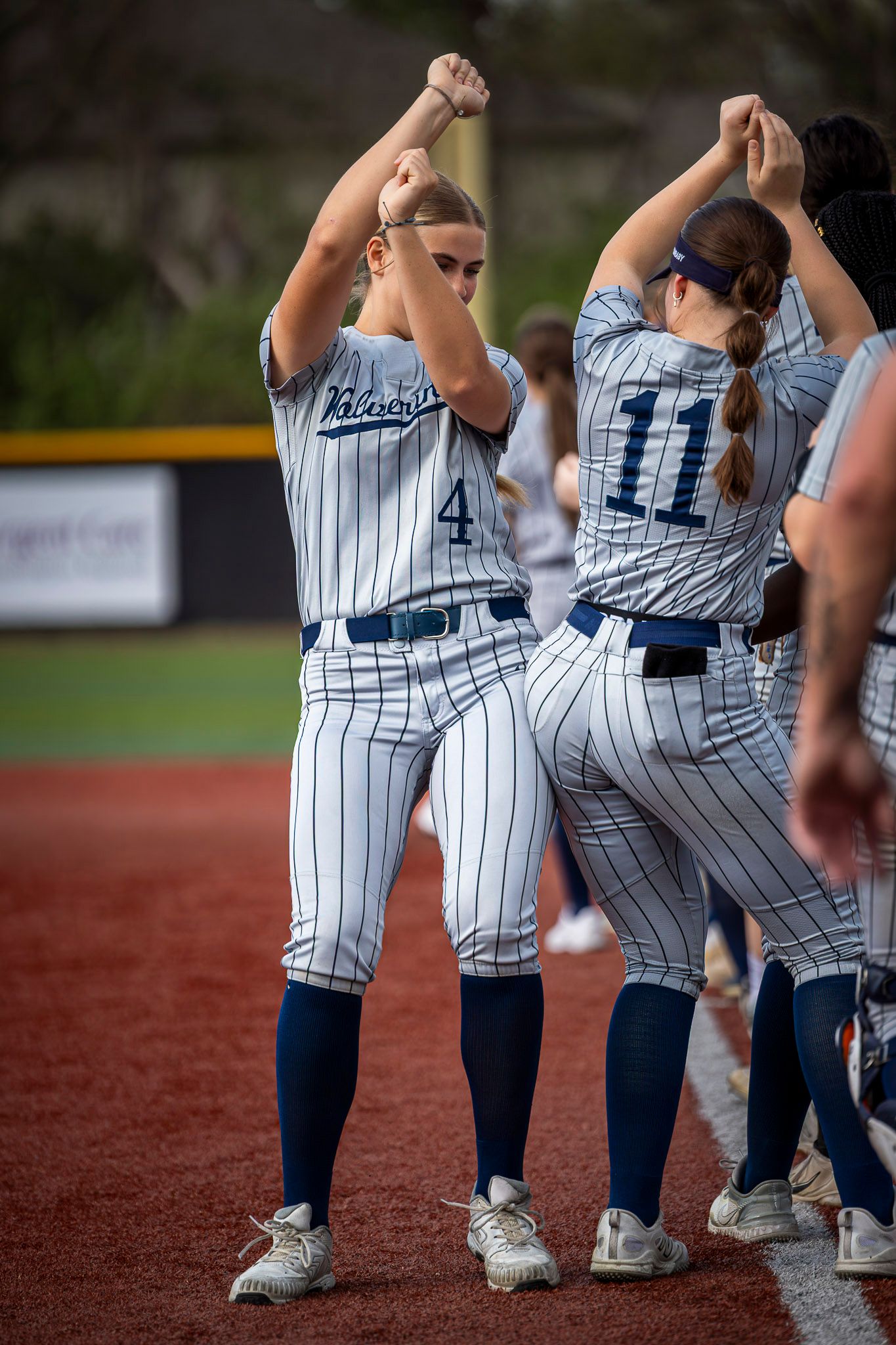 Softball players in white pinstripe uniforms, numbers 4 and 11, raising fists during pre-game ceremony on field.
