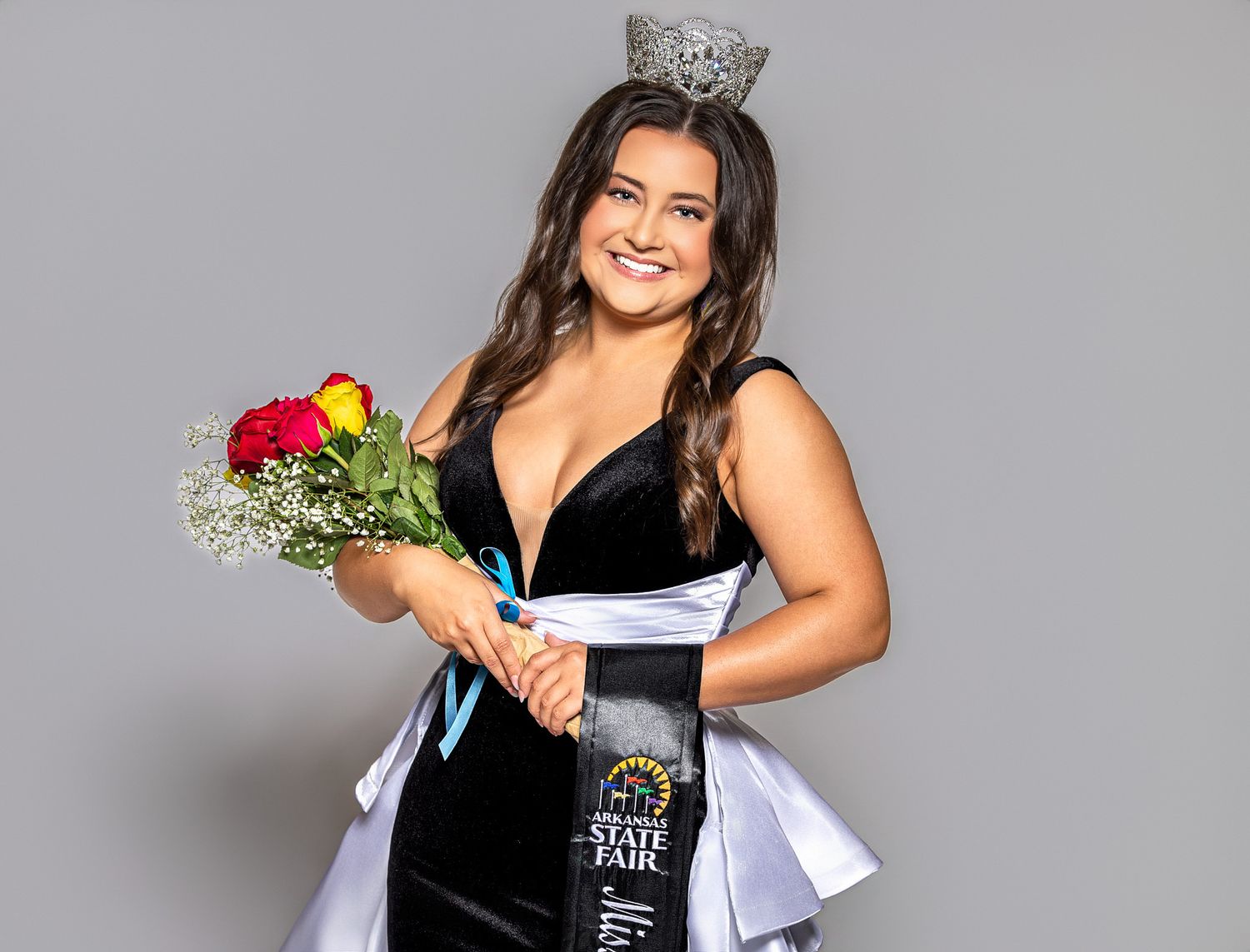 Young woman in black gown wearing a crown and sash, holding red roses, posing as a state fair queen.
