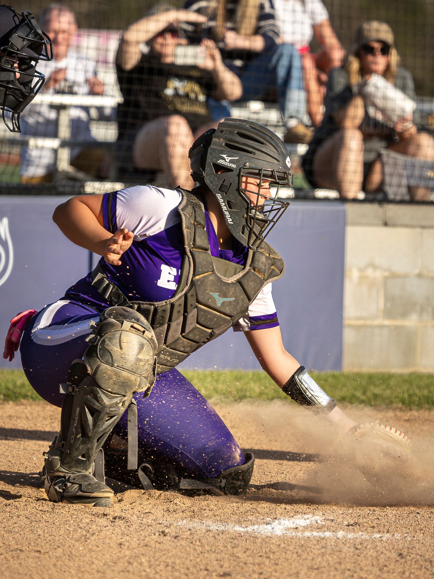 Softball catcher in purple uniform, helmet and chest protector crouches at home plate as dust kicks up while spectators watch from behind.