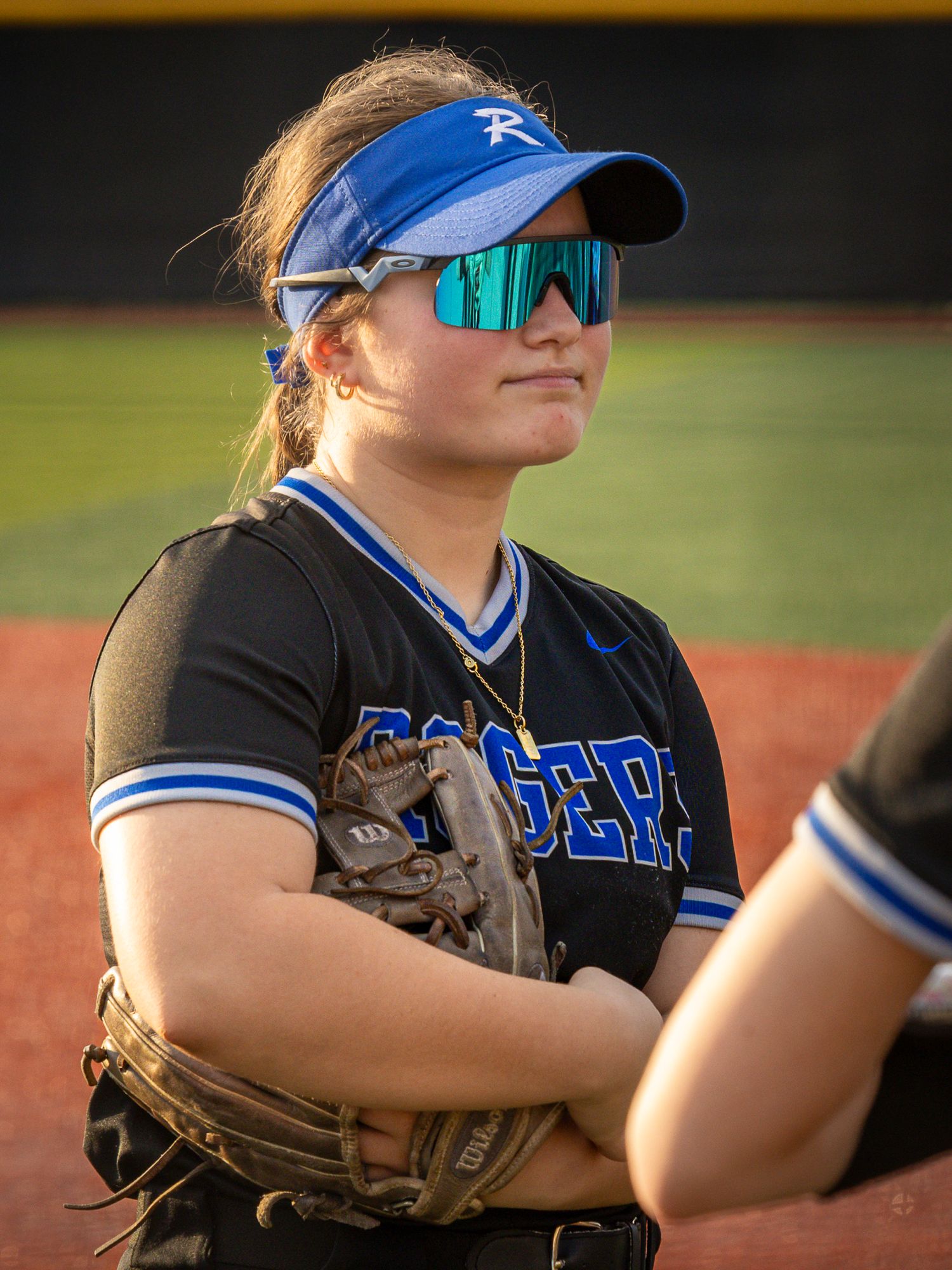 Softball player in a blue visor, teal mirrored sunglasses and black jersey stands with a glove tucked under her arm on the field.