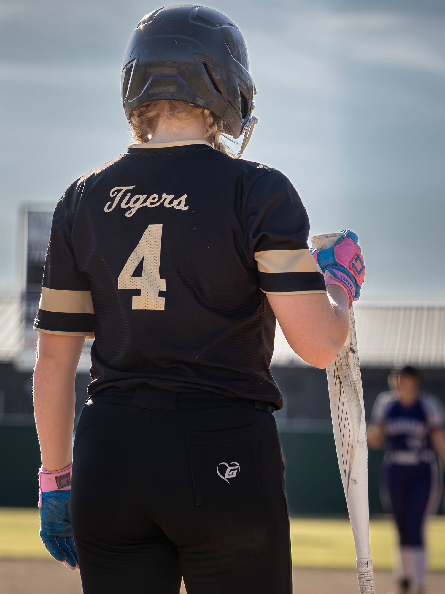 Softball player in a black Tigers jersey number 4 and batting helmet stands holding a bat on the field, seen from behind.