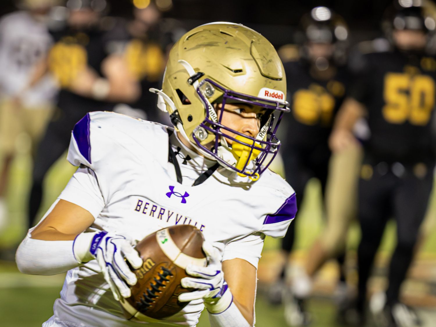 High school football player in a white Berryville jersey and gold helmet carries the ball during a night game as opponents in black look on.