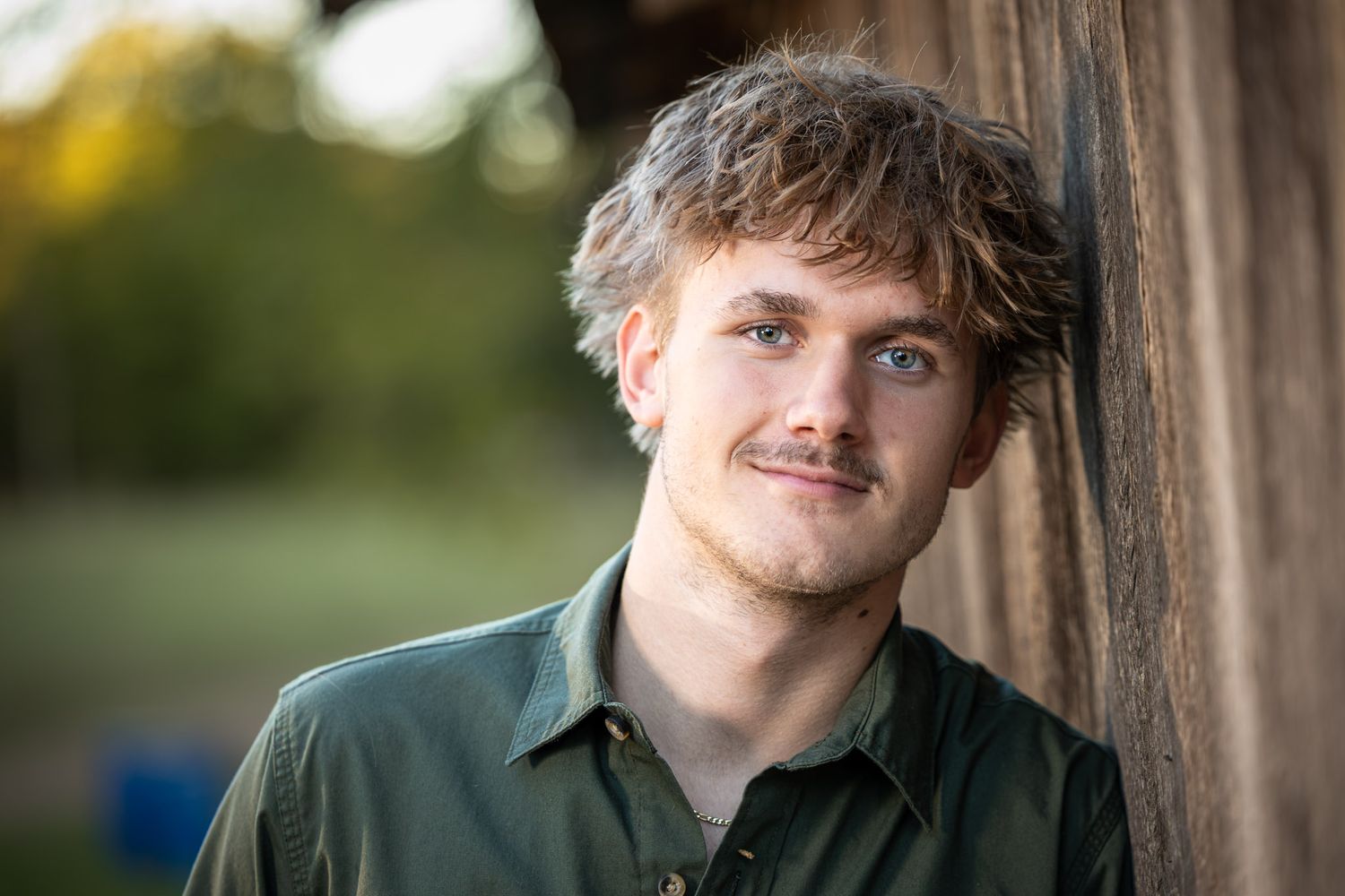 Young man with tousled curly hair and a light mustache leans against a rustic wooden wall, wearing an olive green button-up shirt outdoors.