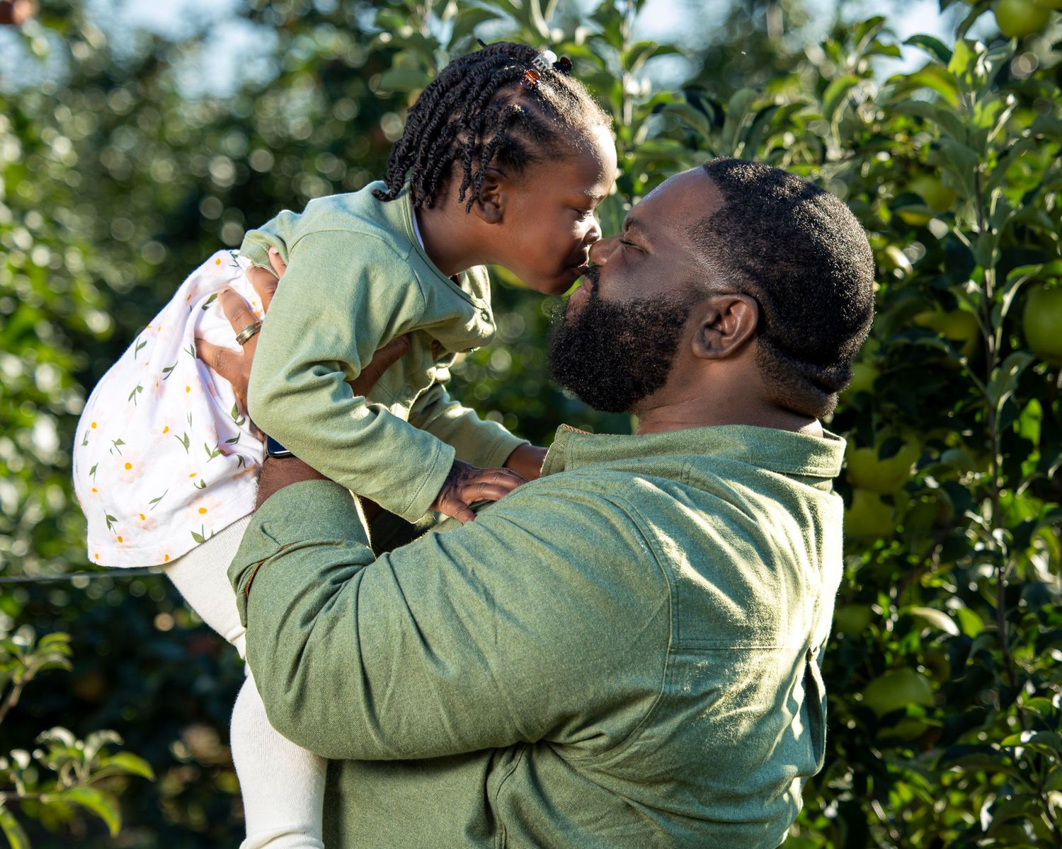 A bearded man lifts a young girl for an Eskimo kiss outdoors among green trees.