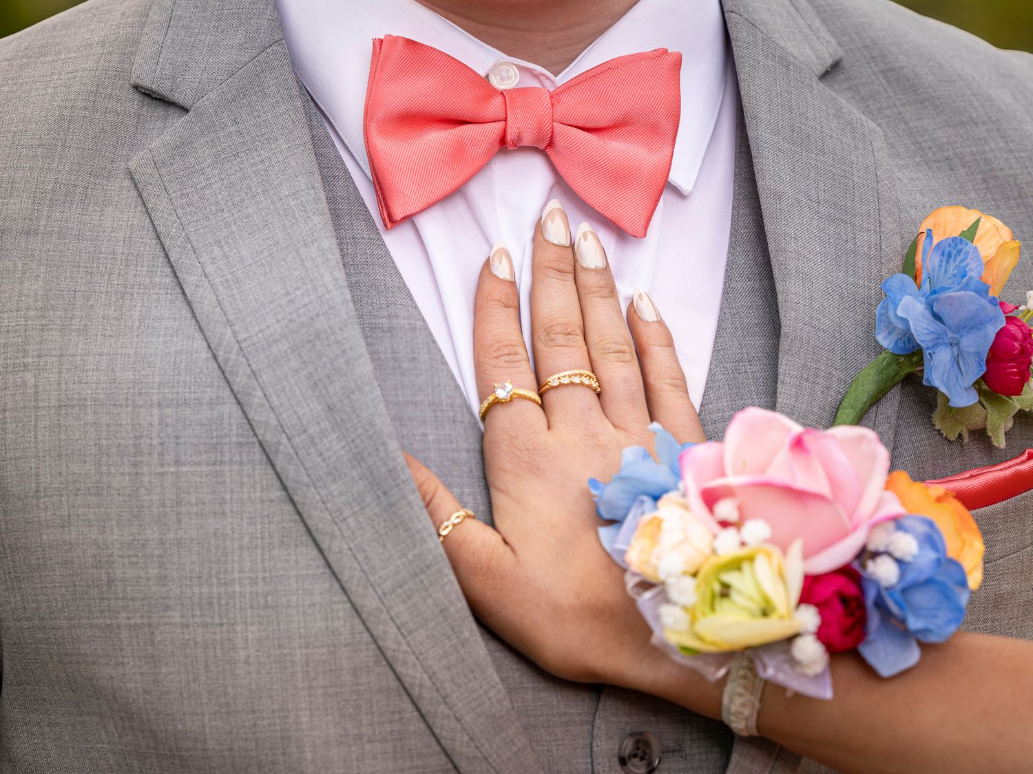 Close-up of a groom in gray suit with coral bow tie and boutonniere, bride's hand with floral wrist corsage resting on his chest.