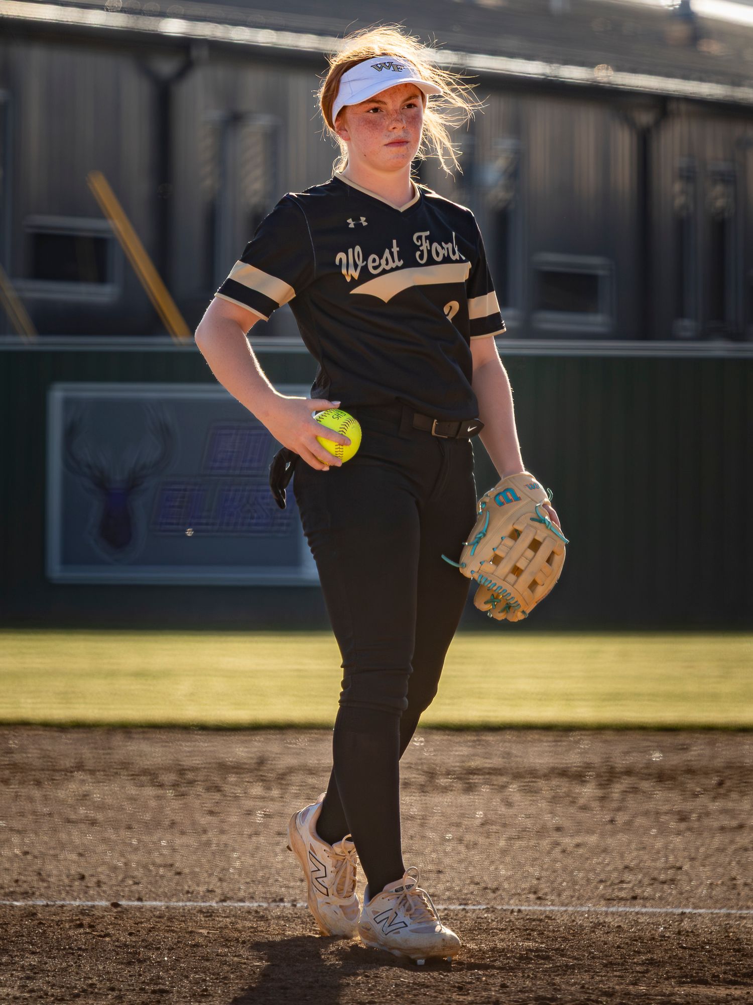 West Fork softball player in a black uniform and white visor holds a yellow softball and mitt while standing on the infield dirt.