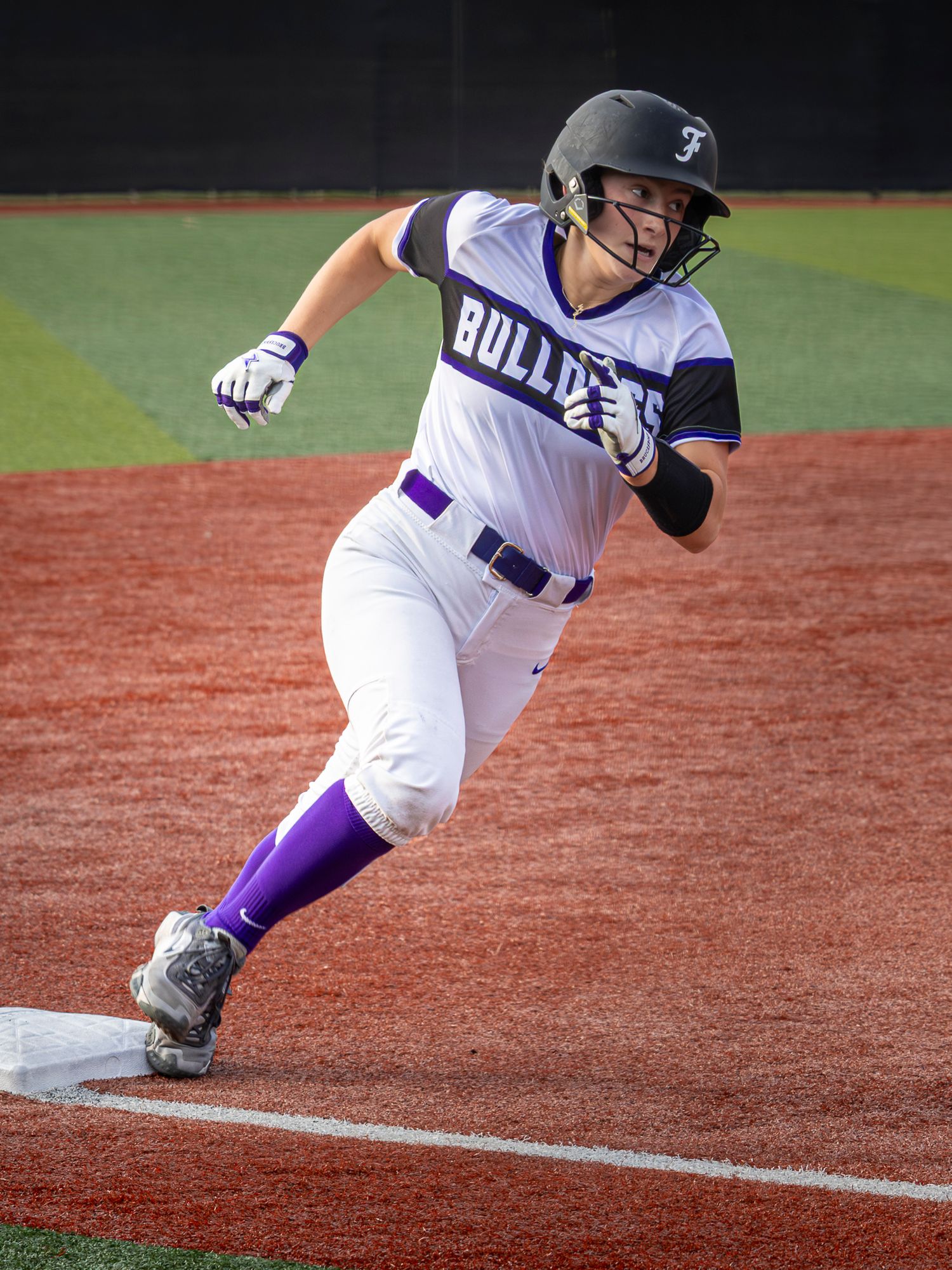 Bulldogs softball player in a white and purple uniform and batting helmet rounds third base while running on the dirt infield.