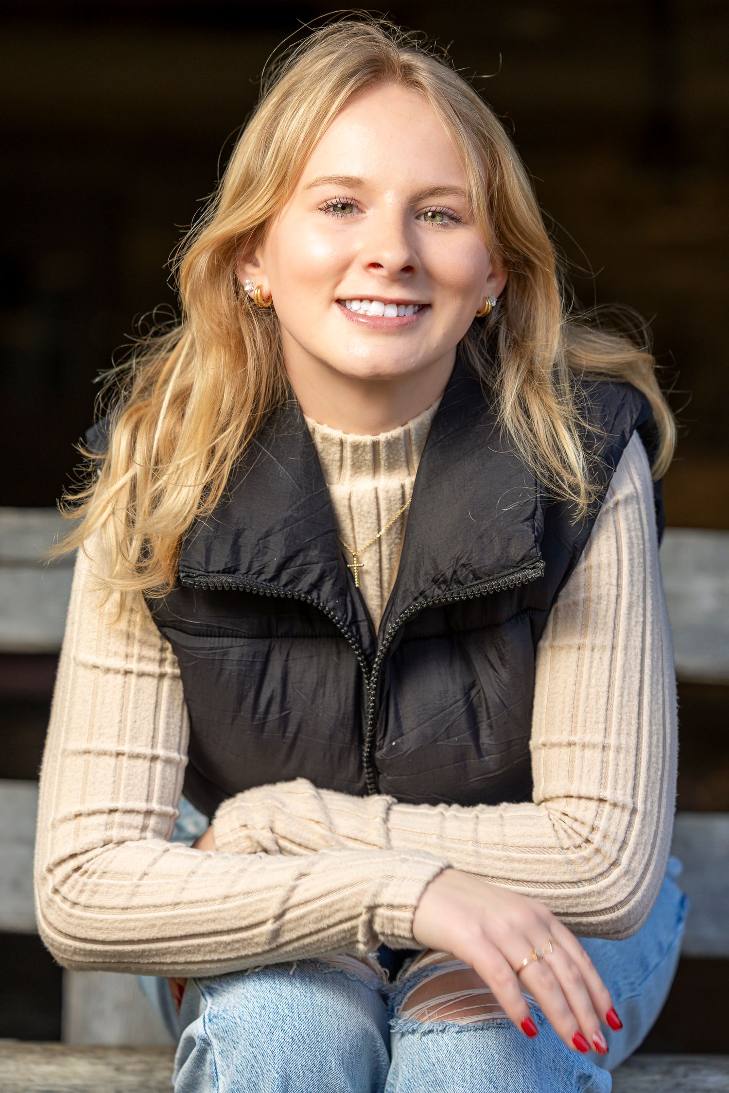 Smiling young blonde woman in black vest and beige sweater sitting outdoors with arms crossed, red nails.