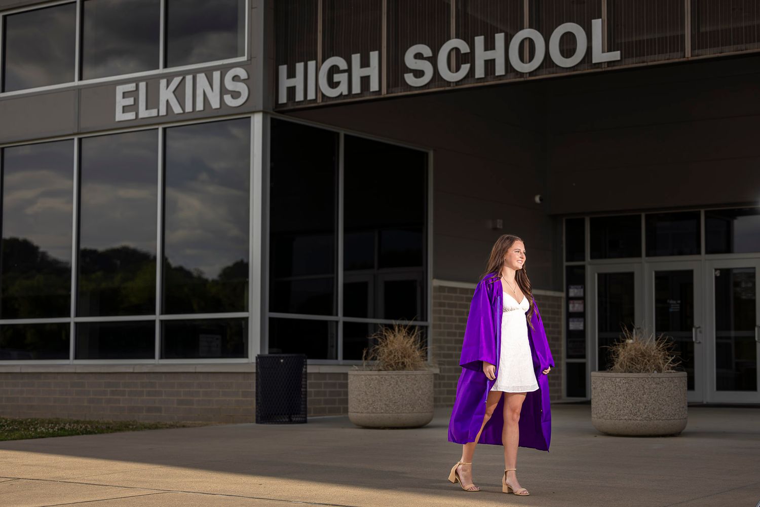 Senior high school graduate in a purple cap and gown poses outside Elkins High School on a sunny day.