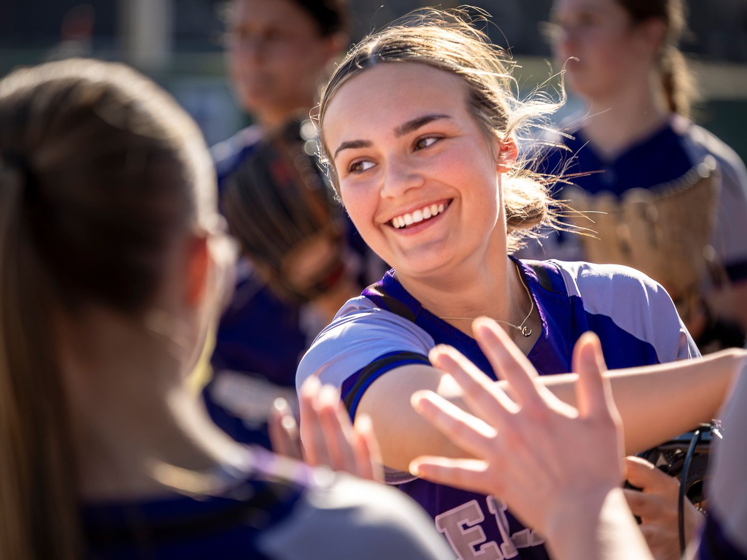 Smiling softball player in a purple uniform shares a joyful moment with teammates, reaching out for high-fives after a game.