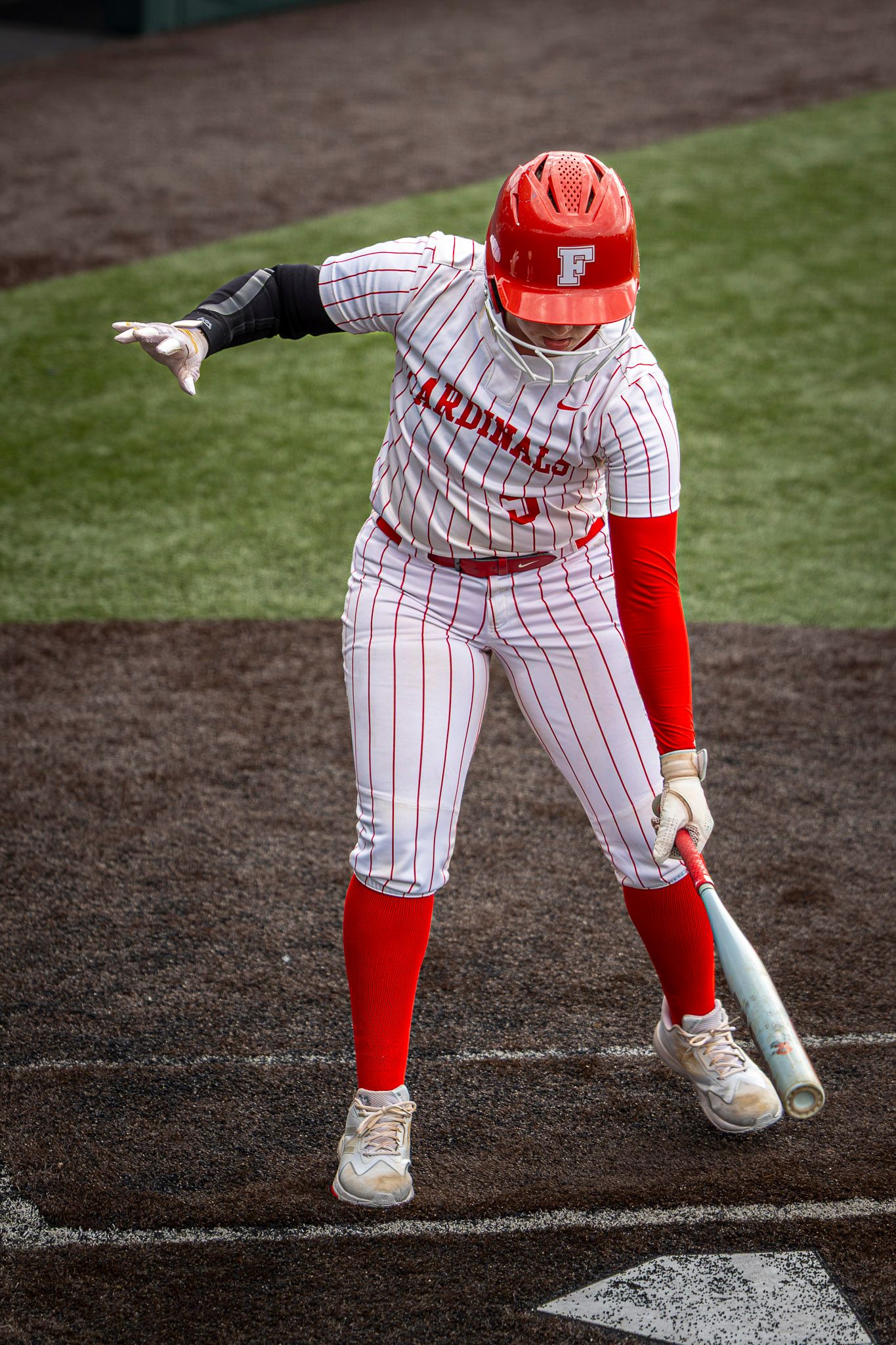 Softball batter in a red helmet and white pinstriped Cardinals uniform stands at home plate holding a bat and pointing with one arm.