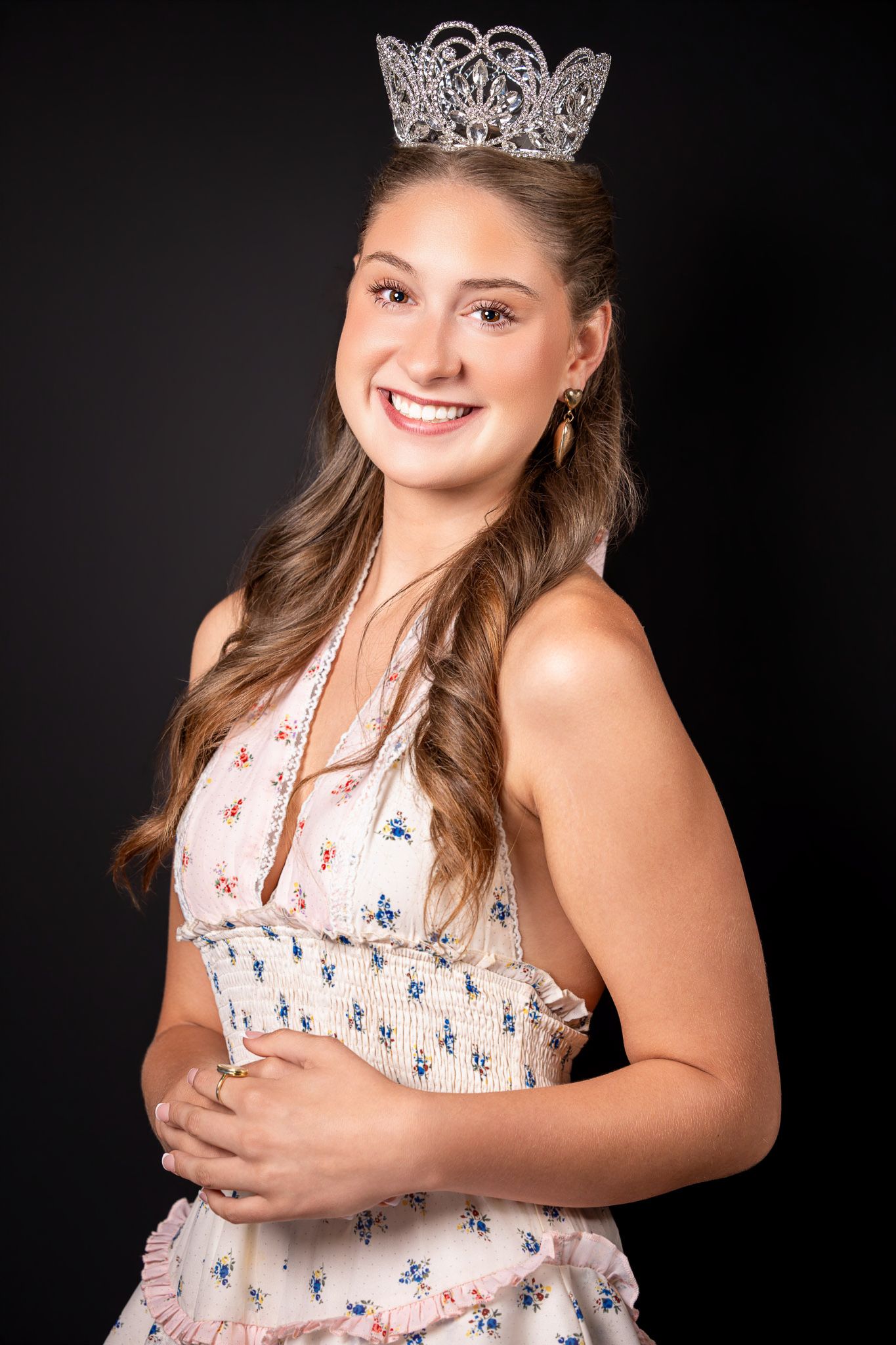 Smiling young woman wearing a sparkling tiara crown and floral dress posing against a dark background.