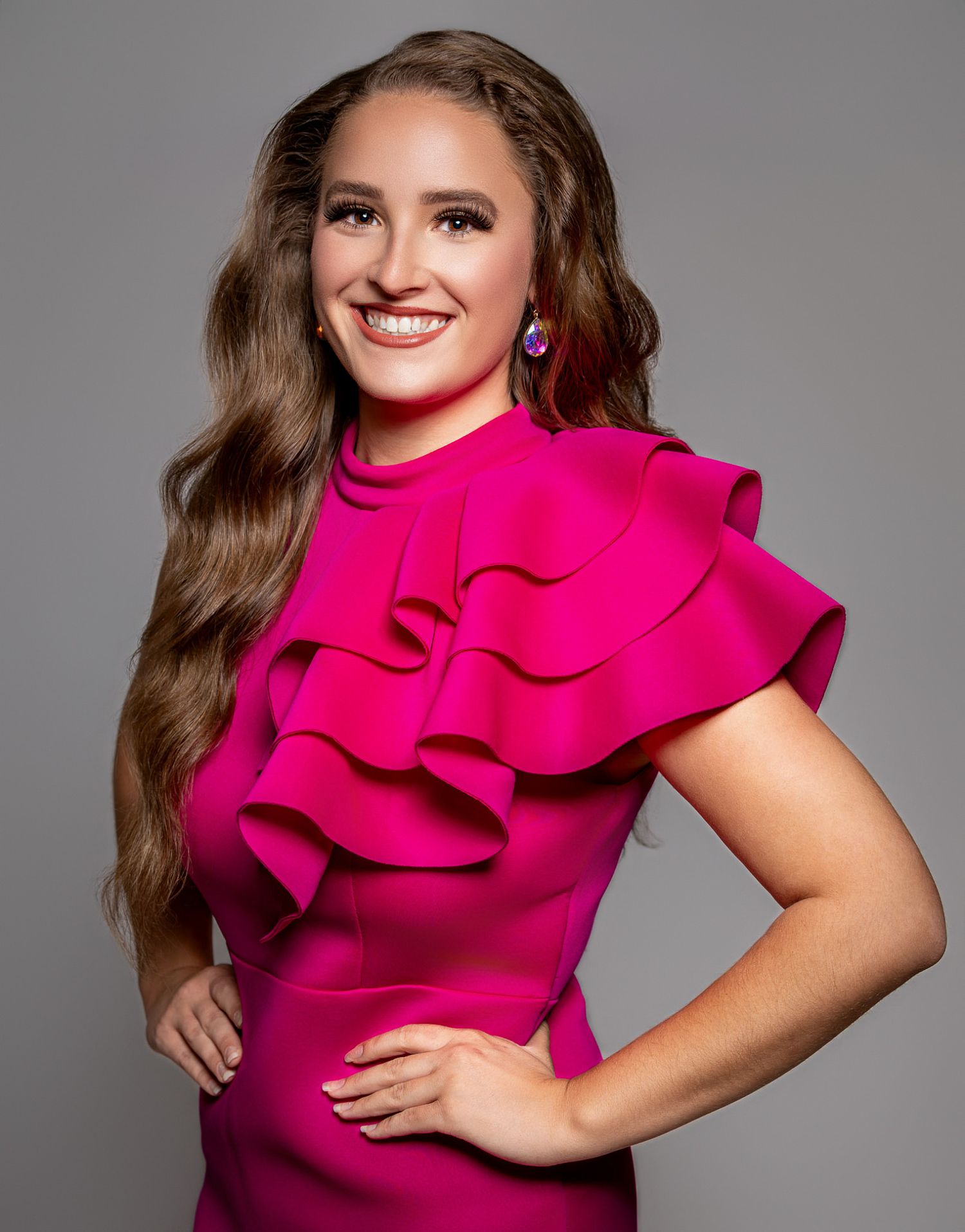 Young woman smiling in a hot pink ruffled dress with blue earrings, posing confidently against a gray background.