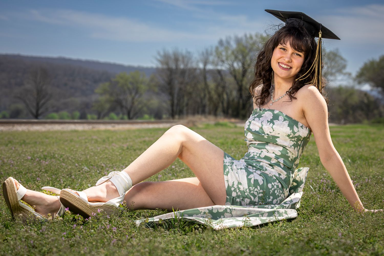 A young woman in a floral dress and graduation cap sitting on a blanket in a grassy field.