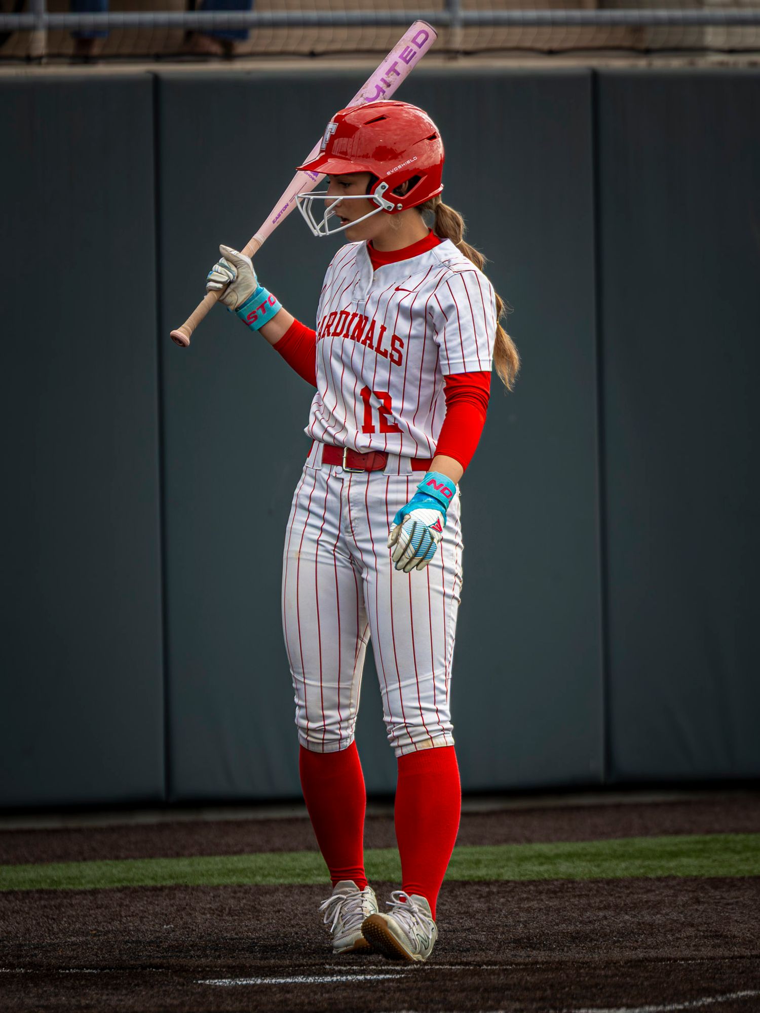 Softball batter in a red Cardinals uniform and helmet stands at home plate holding a pink bat on her shoulder.