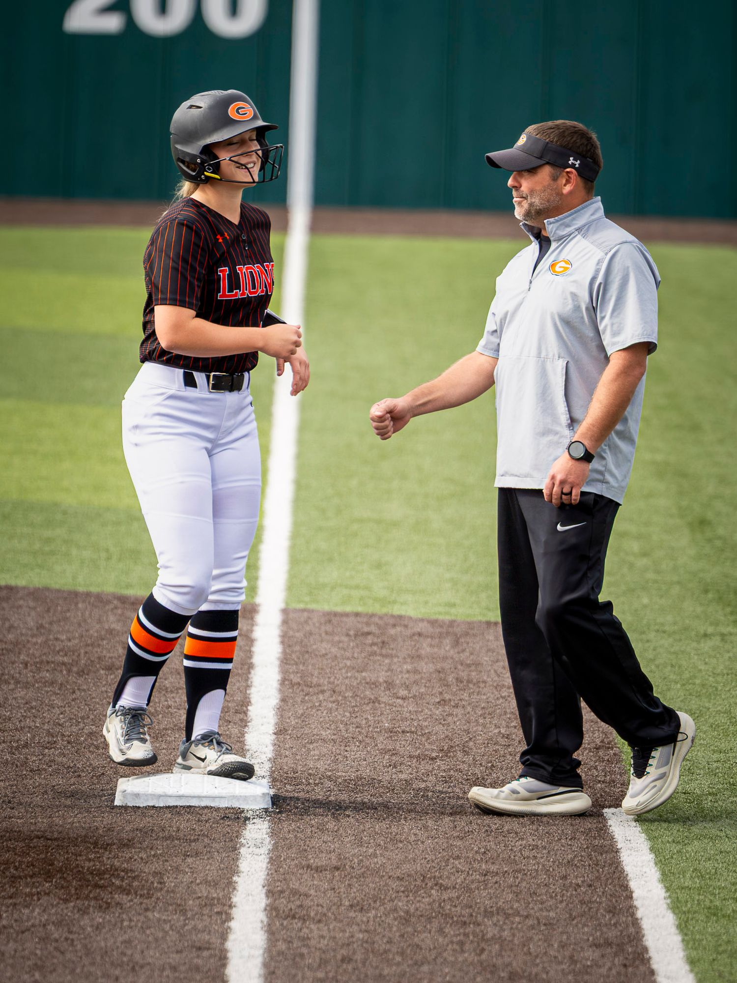 Smiling softball player in a helmet and striped socks stands on third base while a coach in a gray polo and cap talks to her.