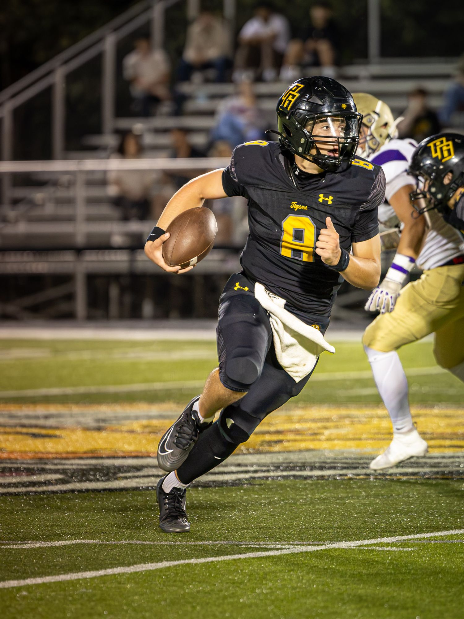 Football quarterback in a black and gold uniform with number 9 runs while gripping the ball during a night game as a defender pursues.