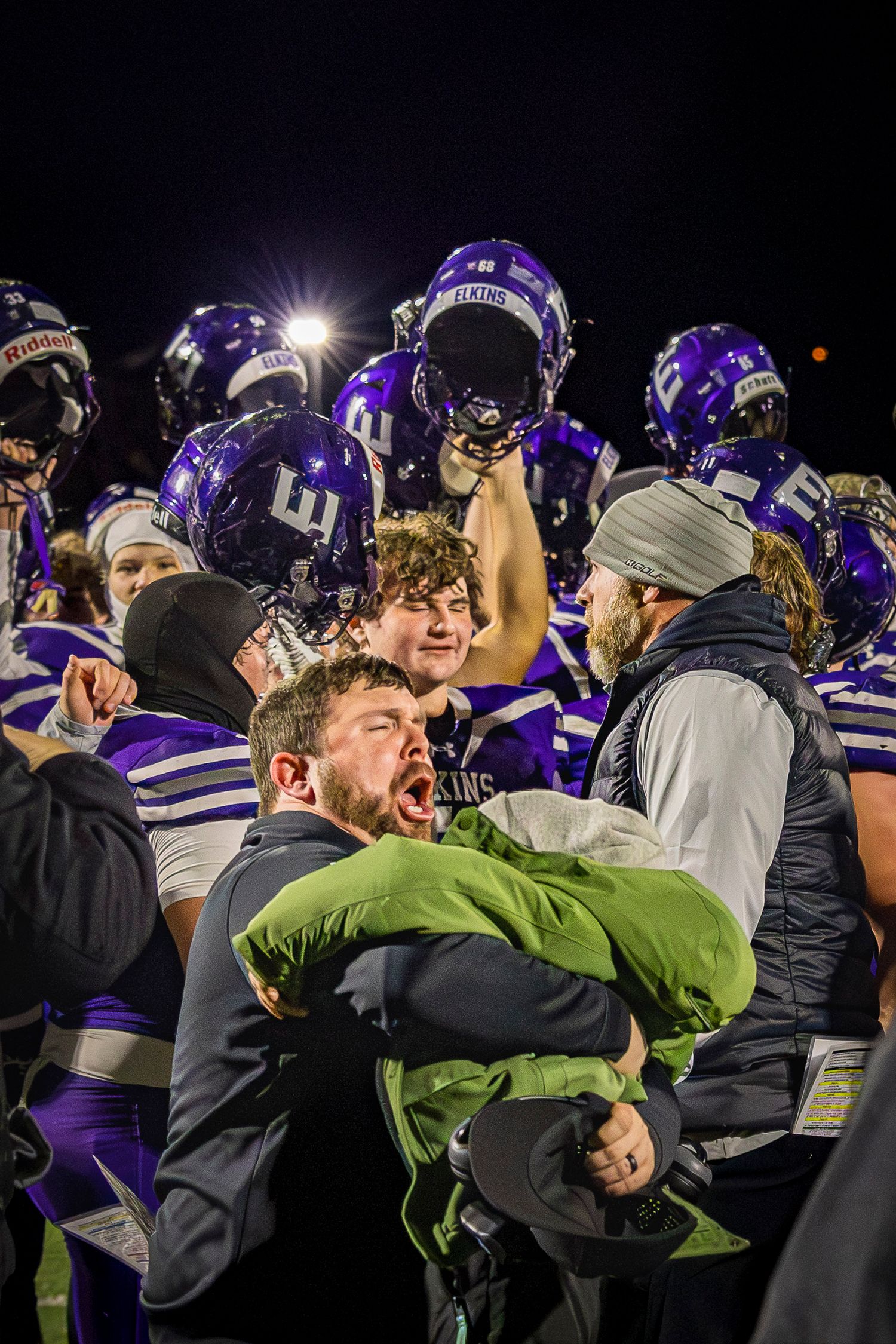 Football team celebrates victory with coaches and players hugging and raising helmets under stadium lights in purple uniforms.