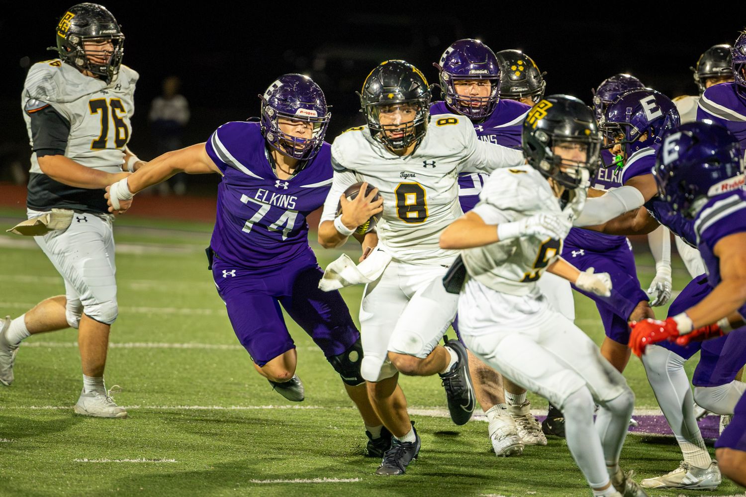 High school football player in a white uniform with number 8 carries the ball through defenders in purple jerseys during a night game.