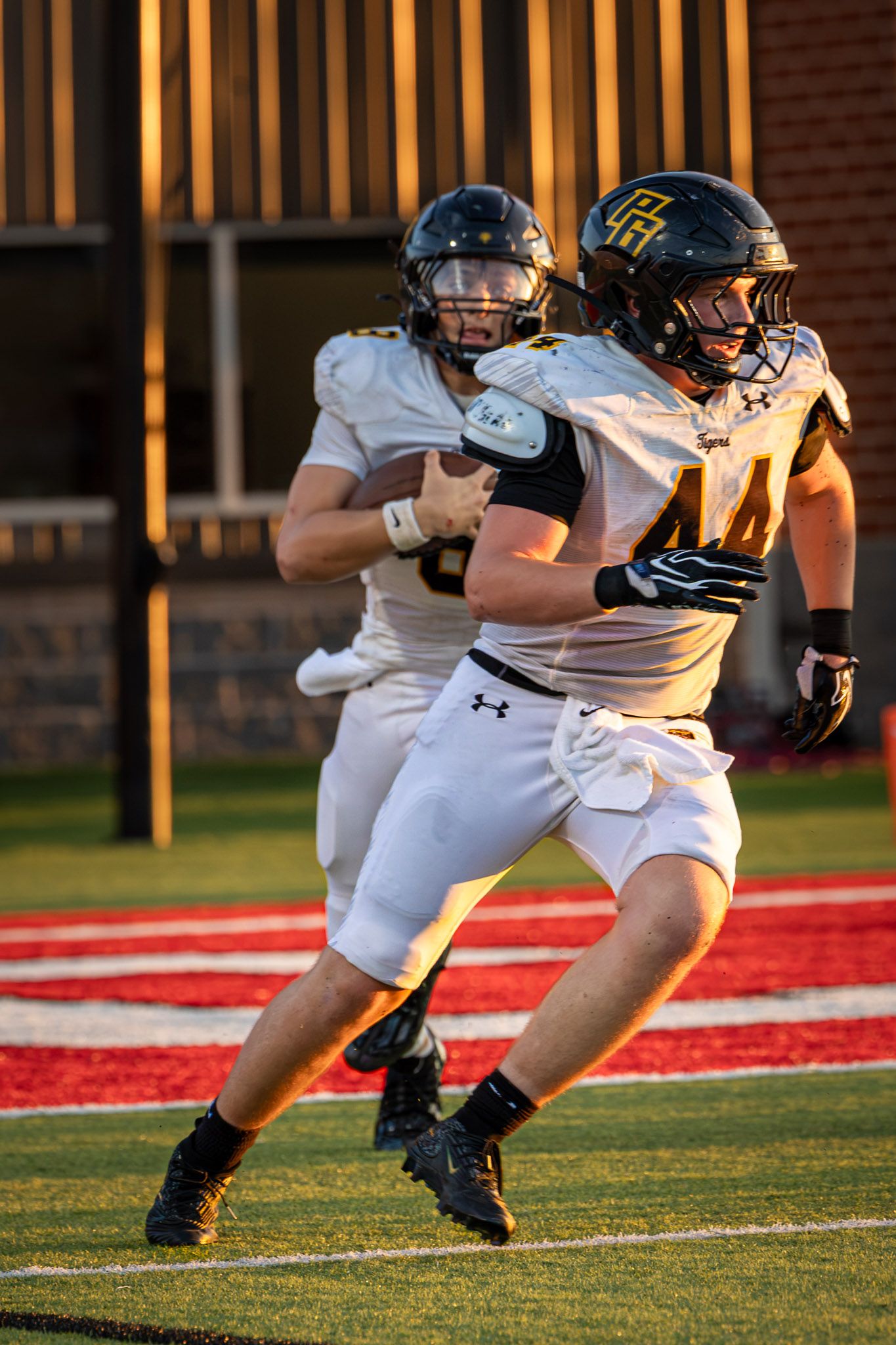 Two football players in white jerseys, black helmets and gold-trimmed uniforms run across a red end zone during an evening game.