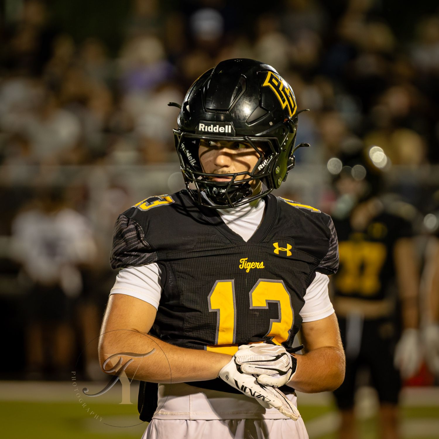 Football player wearing black Tigers jersey number 13 and Riddell helmet stands on field during night game.