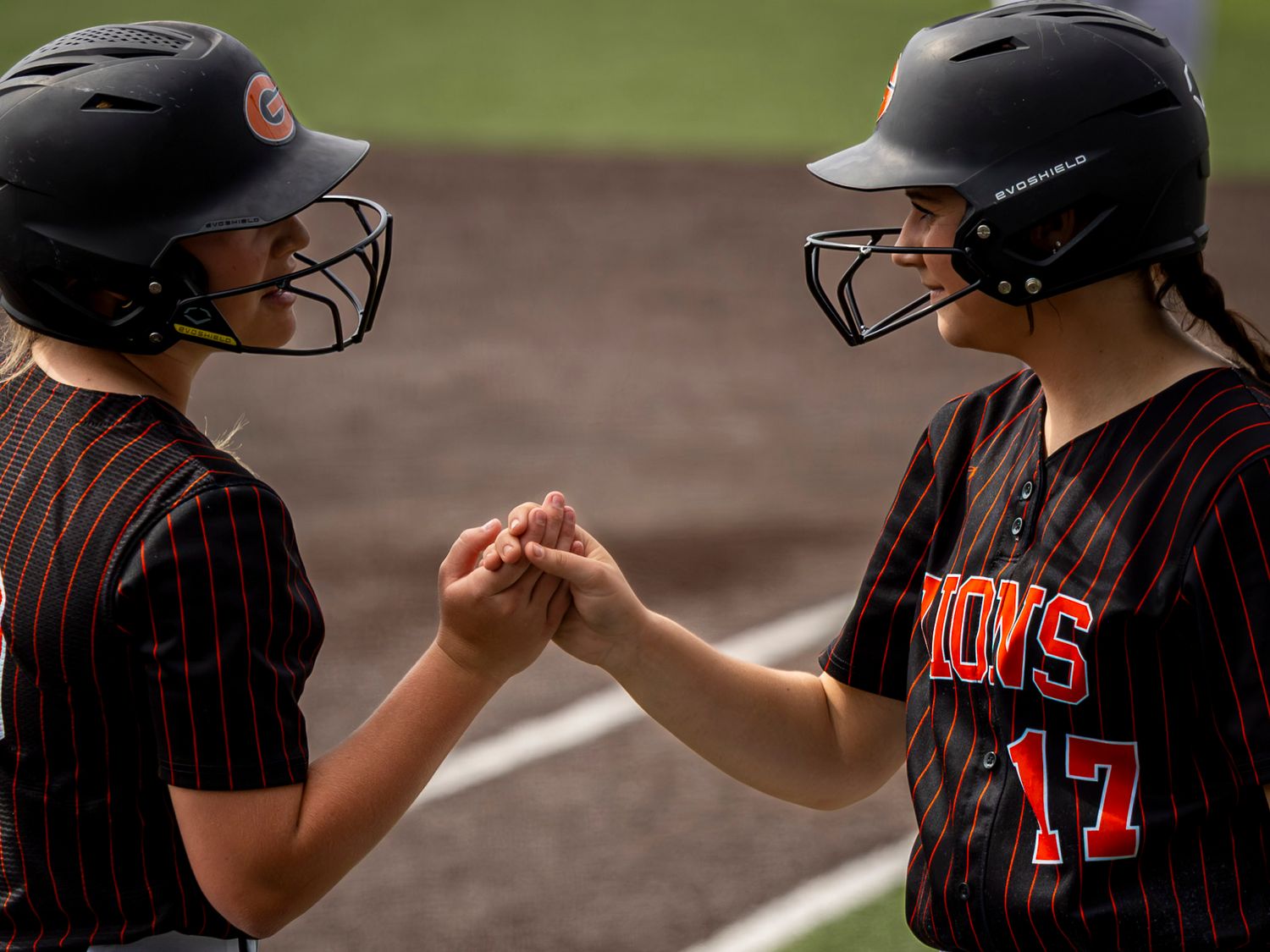 Two softball players in black batting helmets with face guards and pinstriped jerseys share a fist bump on the field during a game.