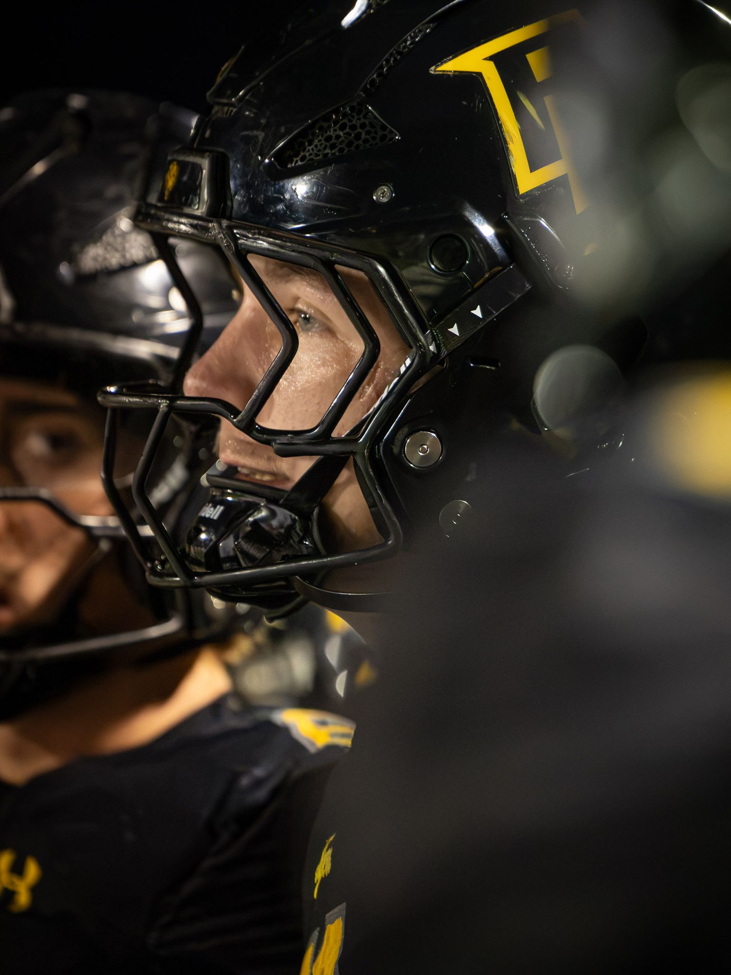 Close-up of a football player in a black helmet with yellow accents and a facemask, standing beside a teammate under dark stadium lighting.