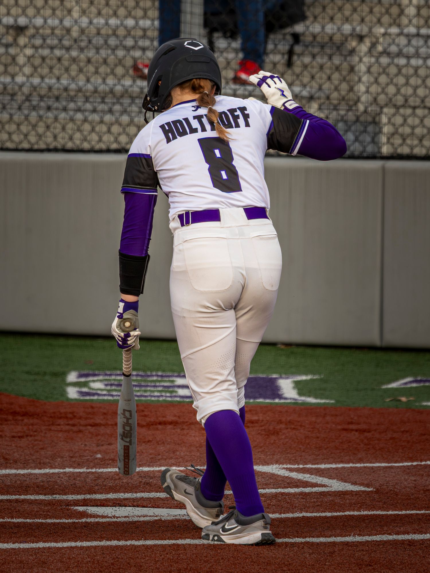 Softball batter in a white jersey labeled Holtyoff number 8 with purple sleeves and socks walks toward the plate holding a bat.