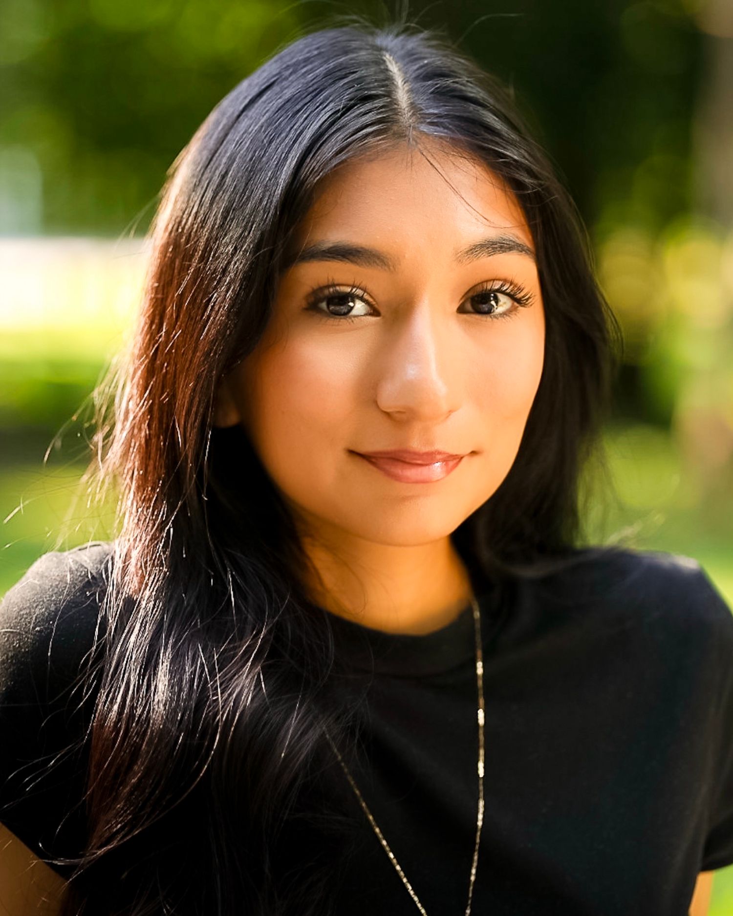 Young woman with long dark hair, a black top and a thin gold necklace smiles softly in an outdoor portrait with a blurred green background.
