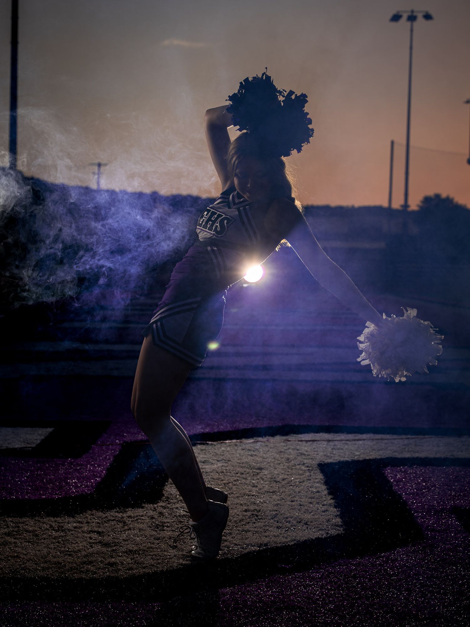 Cheerleader with pom-poms performs dramatic pose at dusk with smoke and lens flare on a track field.