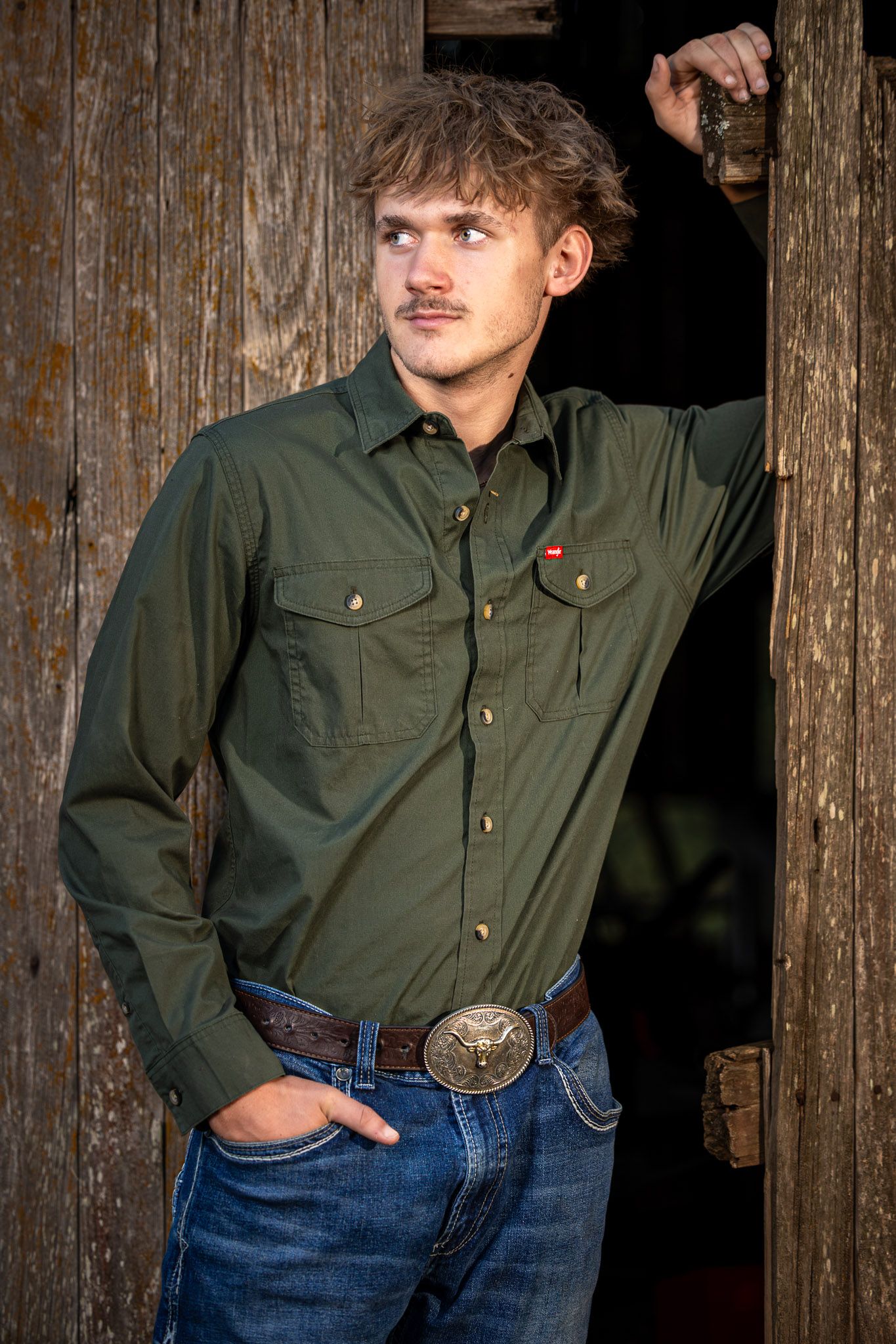 Young man in an olive green button-up work shirt, blue jeans, and a large western belt buckle leaning against a rustic weathered barn door.
