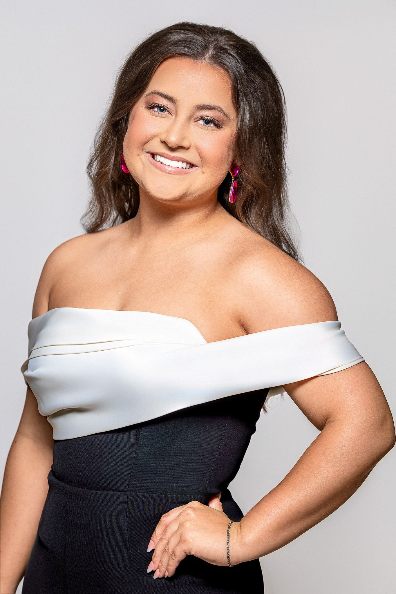 Smiling woman in white off-shoulder top and black outfit with pink earrings poses confidently against gray background.