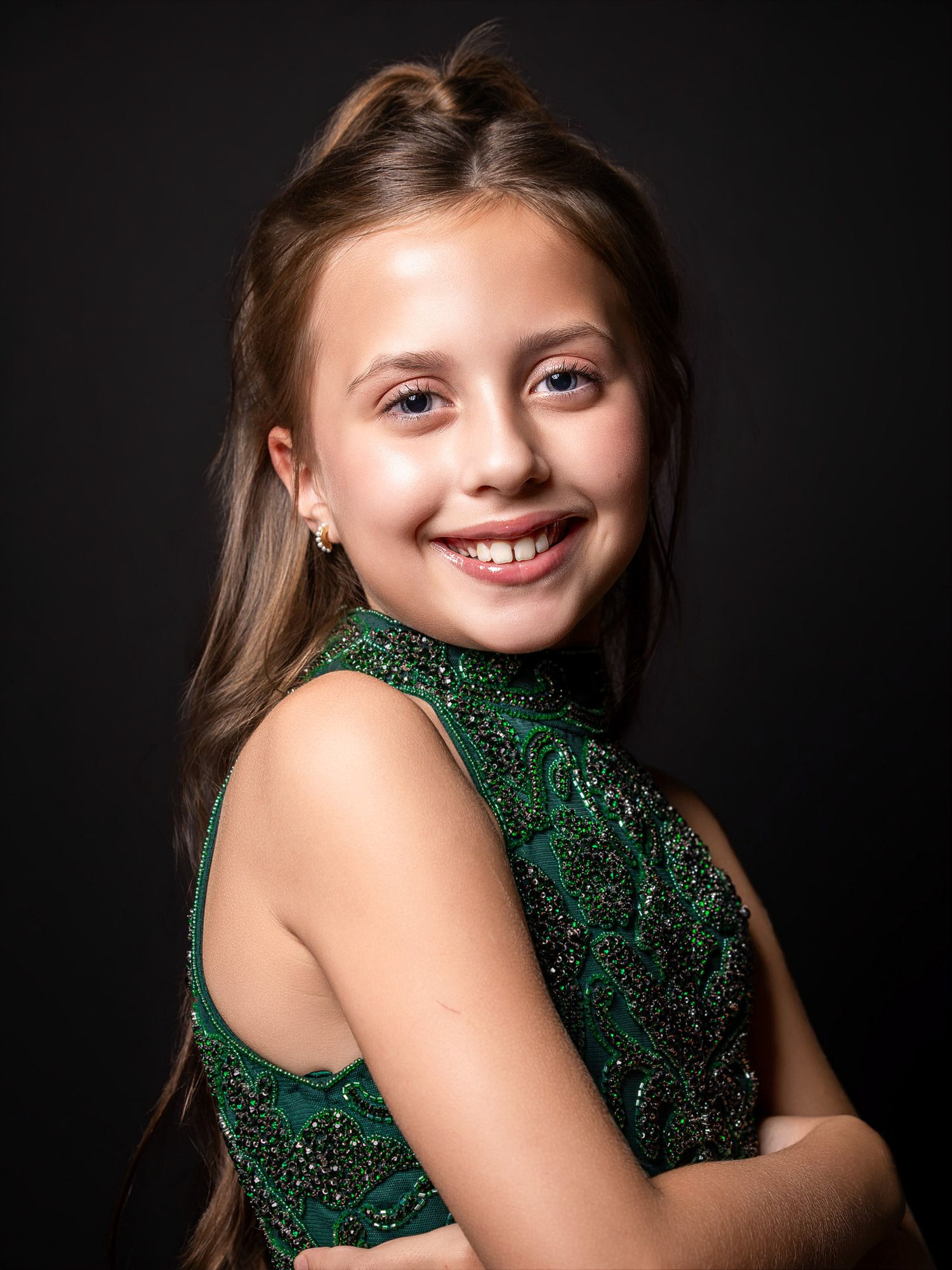 Young girl in a green sequin dress smiling over her shoulder against a dark background.