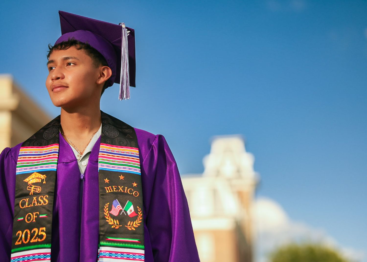 Young Latino graduate in purple cap and gown with Class of 2025 and Mexico stole poses outdoors on a sunny day.