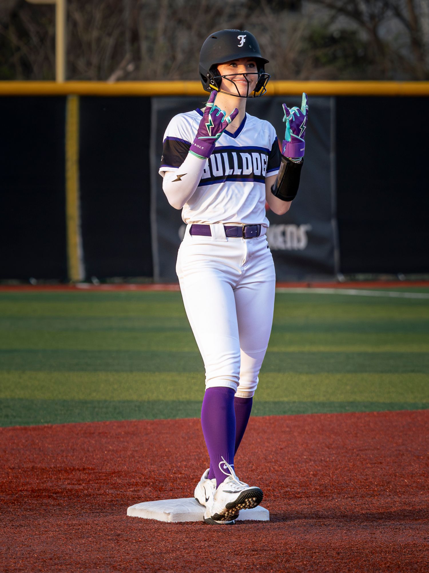 A softball player stands ready on the red dirt infield during a game at dusk.