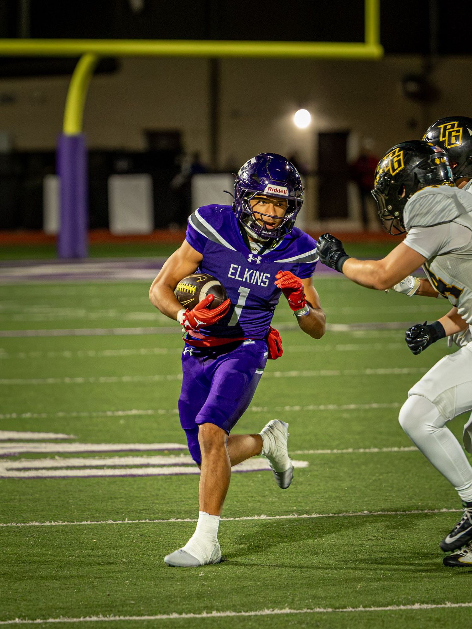 Elkins football player in a purple uniform and helmet carries the ball down the field while a defender in a gold helmet pursues him.