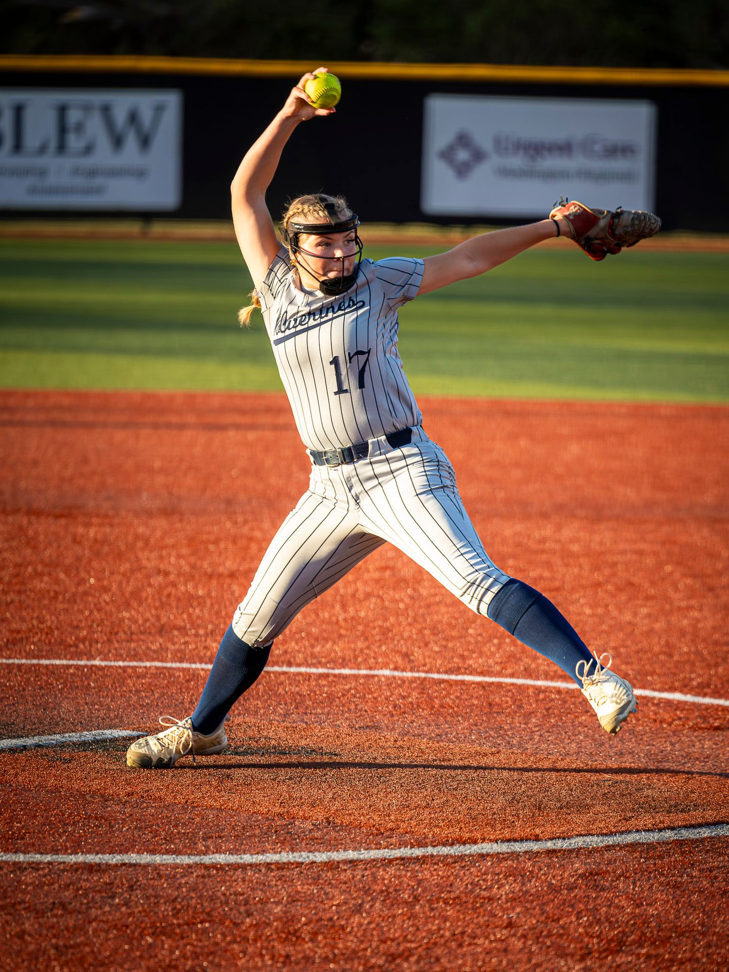 Softball pitcher in a gray pinstriped uniform with number 17 winds up to throw a yellow softball on a red clay infield.
