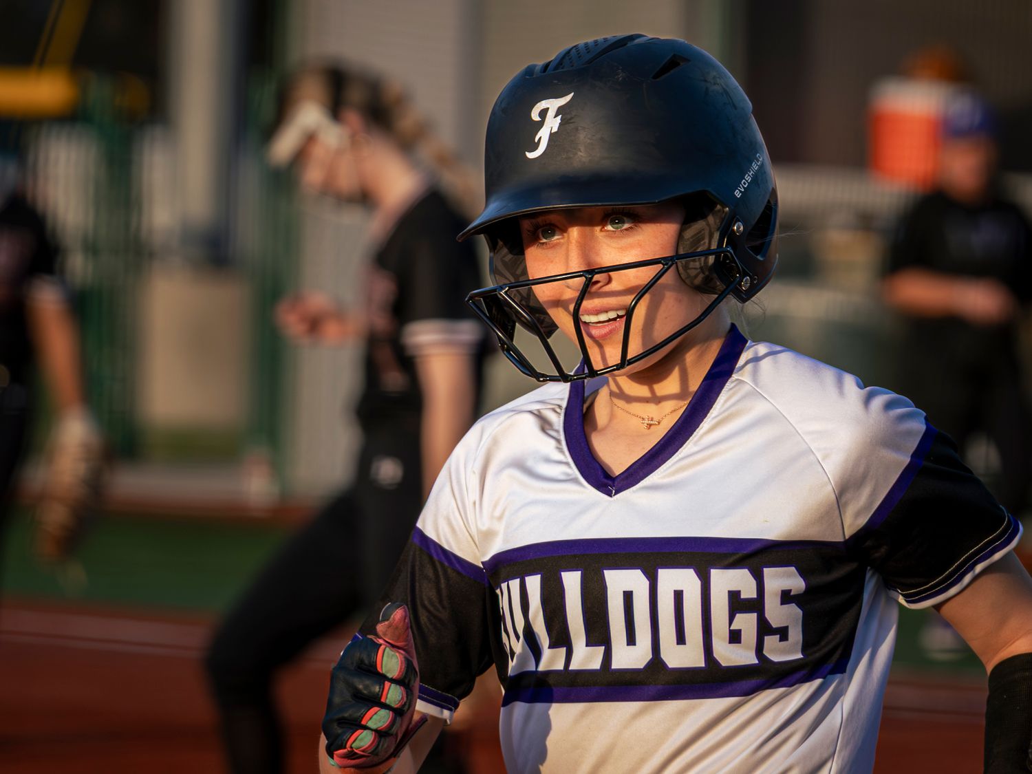 Smiling softball player in a purple and white Bulldogs jersey and batting helmet with face guard stands on the field during an evening game.