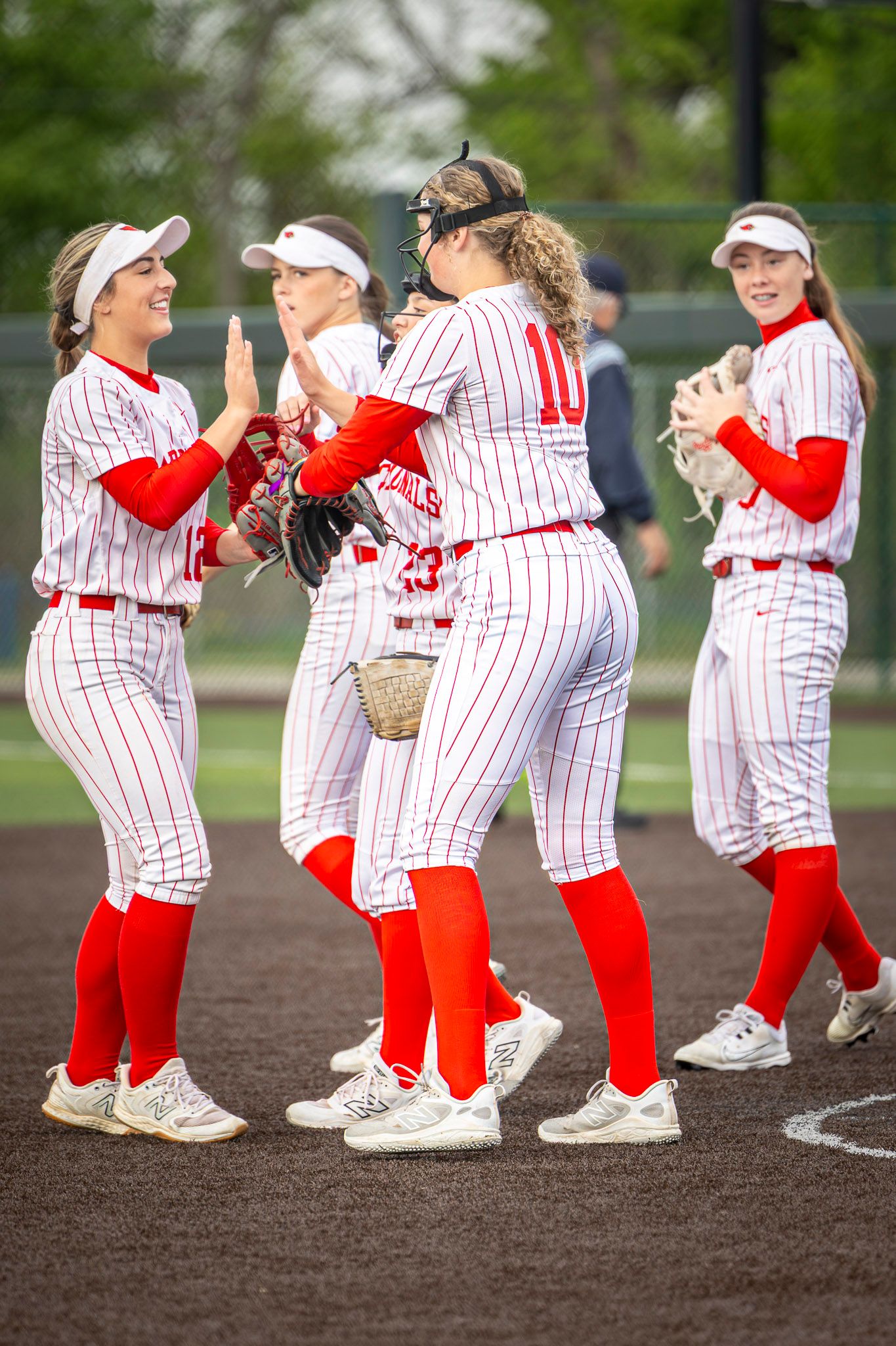 Softball players in red and white pinstriped uniforms celebrate and high-five on the dirt infield during a game.