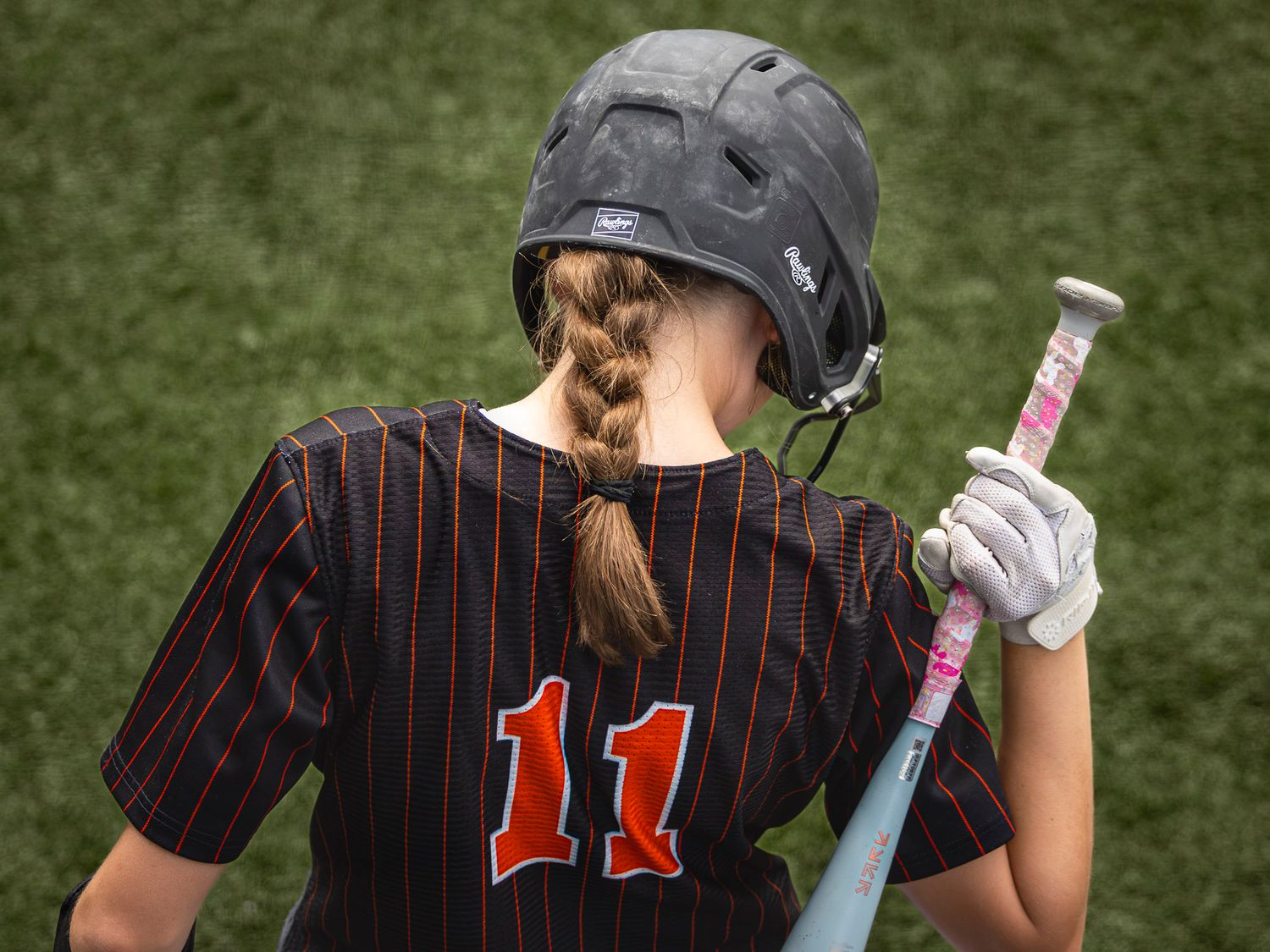 Softball player with a braided ponytail, black helmet and number 11 pinstripe jersey holds a pink bat while walking across the field.