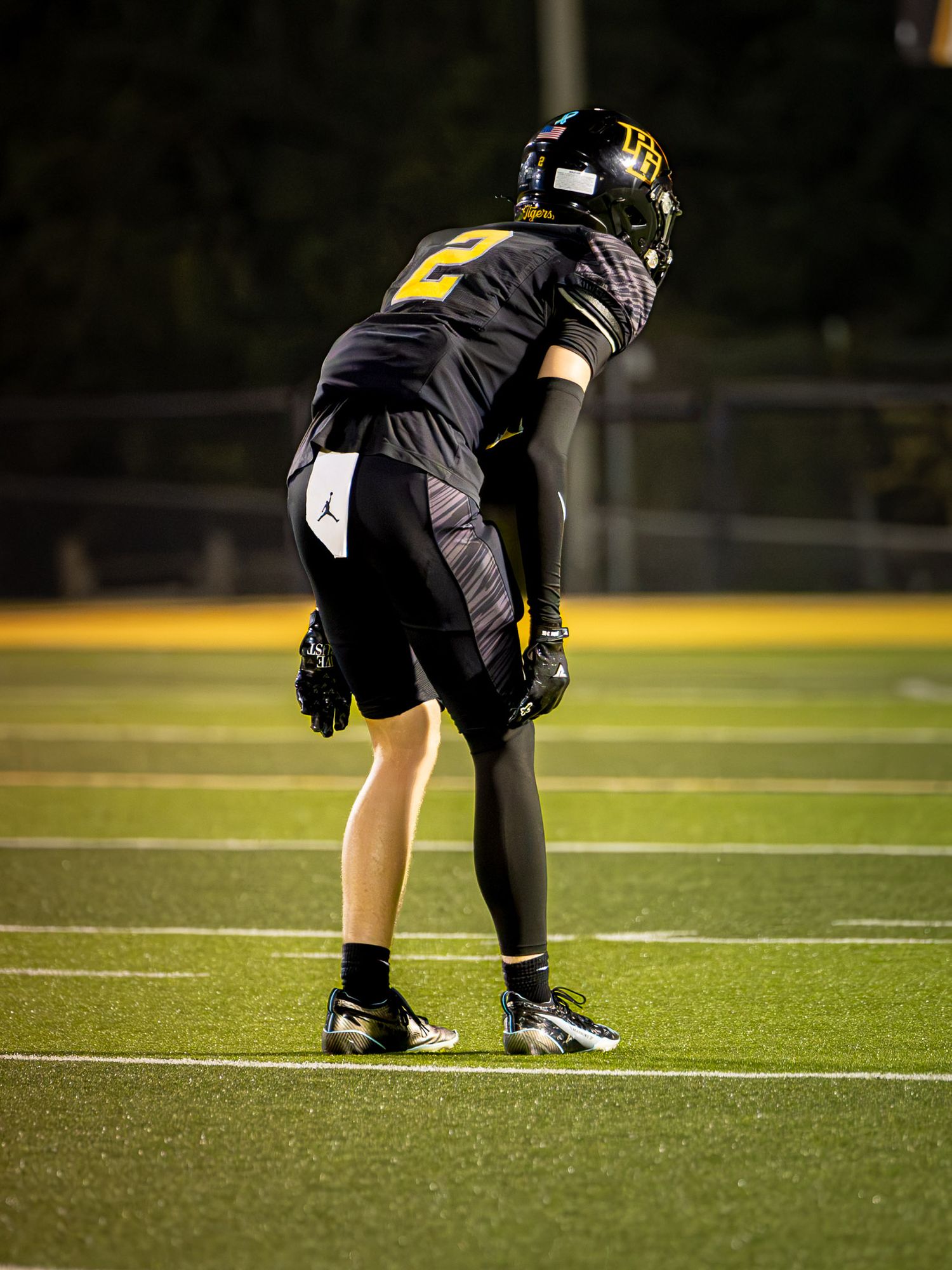 Football player in a black and gold uniform with number 2 crouches in a ready stance on a turf field during a night game.