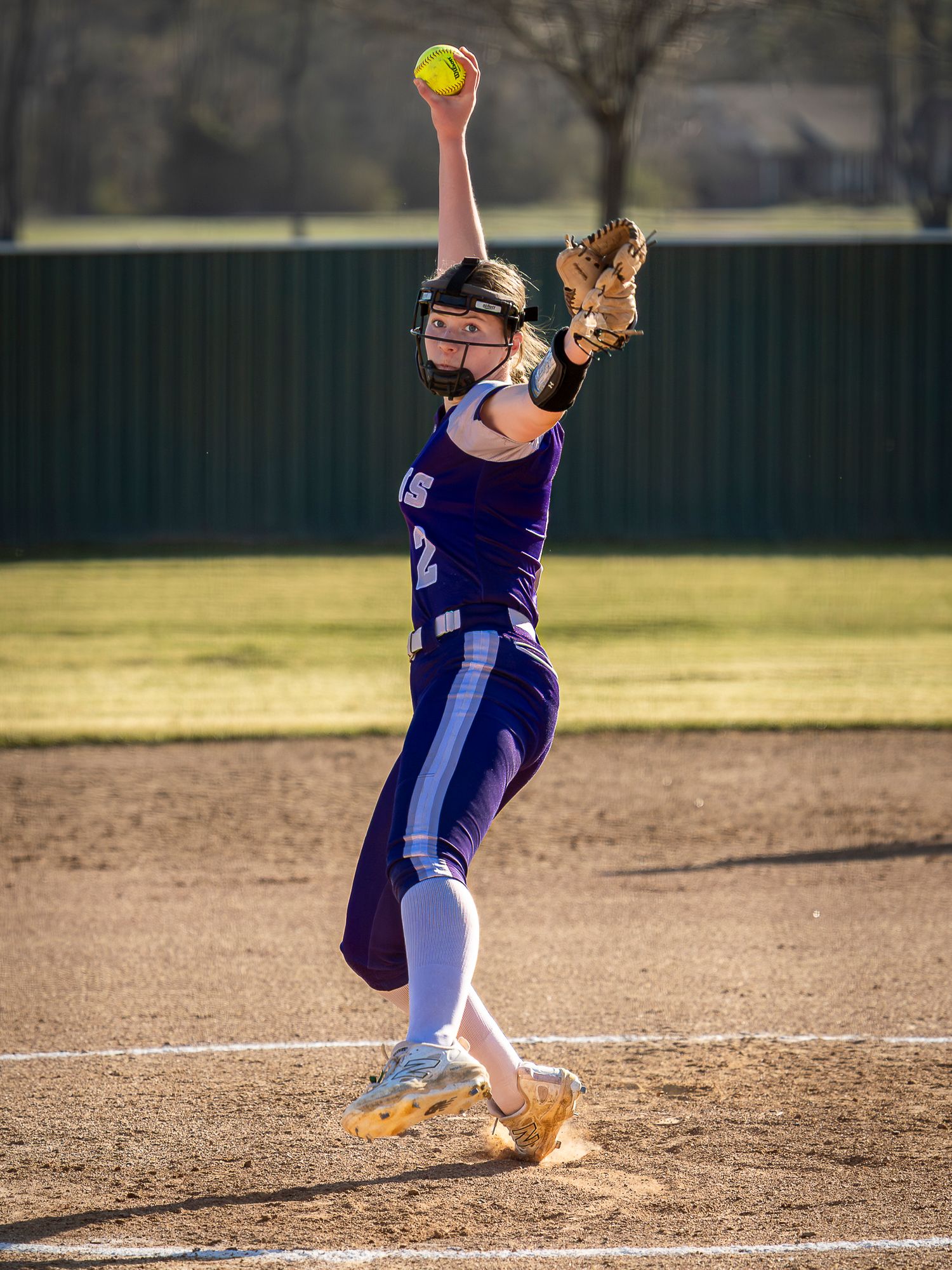 Softball pitcher in a purple uniform and face guard winds up mid-delivery with a yellow softball raised high on a dirt field.