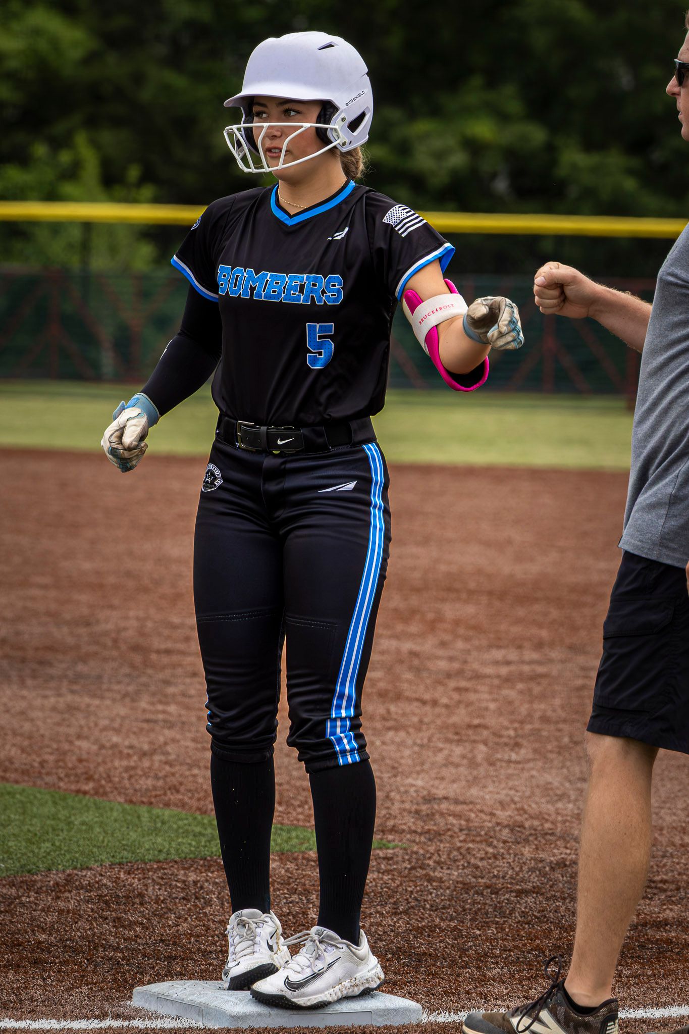 Softball player #5 in Bombers uniform stands on base, wearing white helmet and batting gloves during a game.