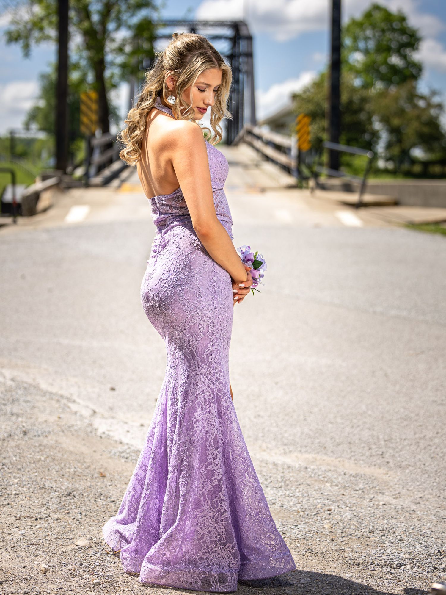 Young woman in elegant lavender lace mermaid prom gown holding bouquet on bridge road.