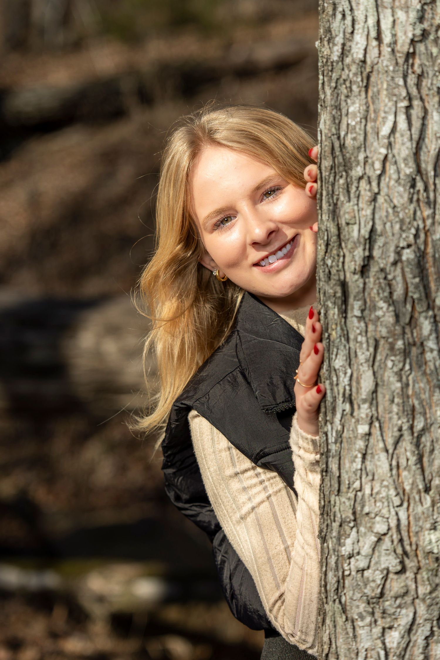 Smiling blonde woman peeking playfully from behind a tree trunk in a sunlit forest wearing a black vest.