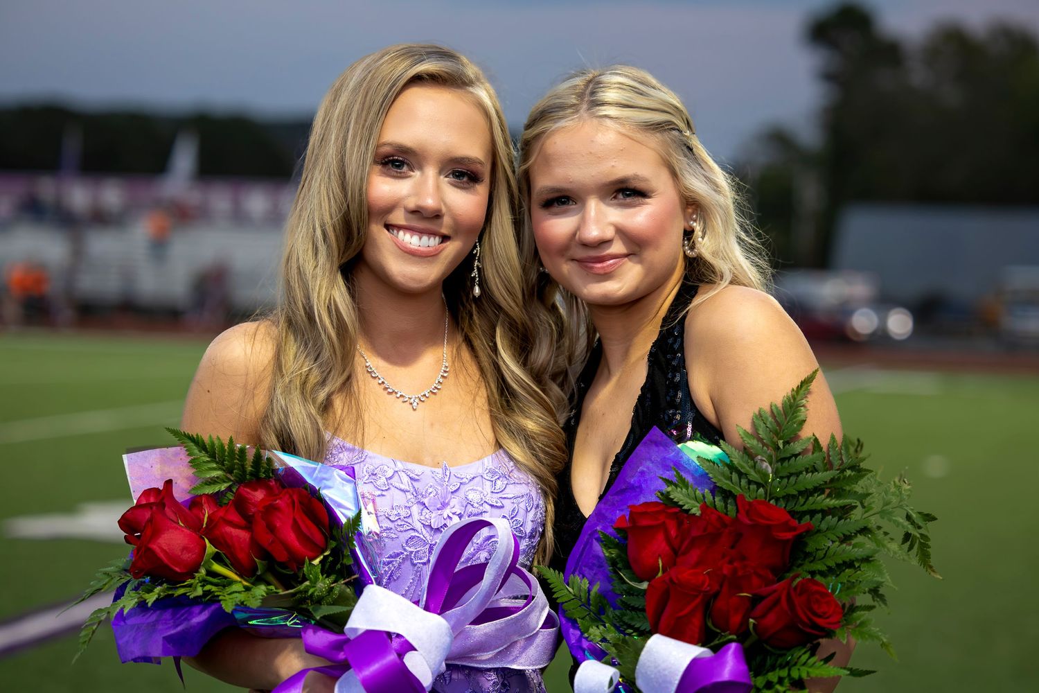 Two smiling young women in formal dresses holding red rose bouquets with purple ribbons on a football field.