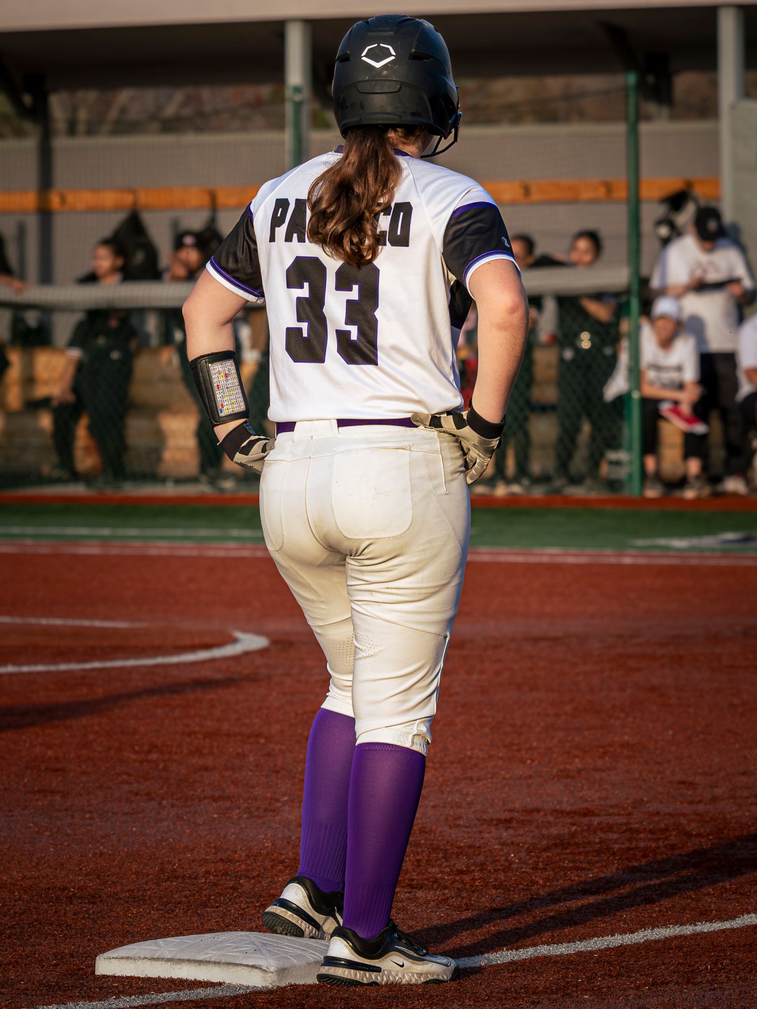 Softball player in white uniform with number 33, purple socks and black batting helmet standing on a base on a red clay infield.